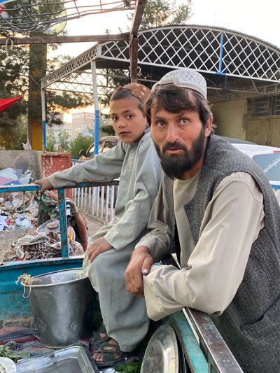 In a candid outdoor shot taken in the city of Kandahar, Afghanistan, a man and a boy sit close together. The man, likely in his late 30s or early 40s, sports a dark beard and a white skullcap, wearing a light-colored shirt with a dark vest. To his left, a young boy, wearing a brown skullcap and a matching light-colored outfit, gazes directly at the camera. Both are positioned within what seems to be the back of a small truck or cart, amidst various items including a large metal pot, a tray, and other miscellaneous objects. In the background, a building with a white, lattice-like awning can be seen, along with hints of trees and a partially visible vehicle. The lighting suggests that the photo was taken either in the early morning or late afternoon.