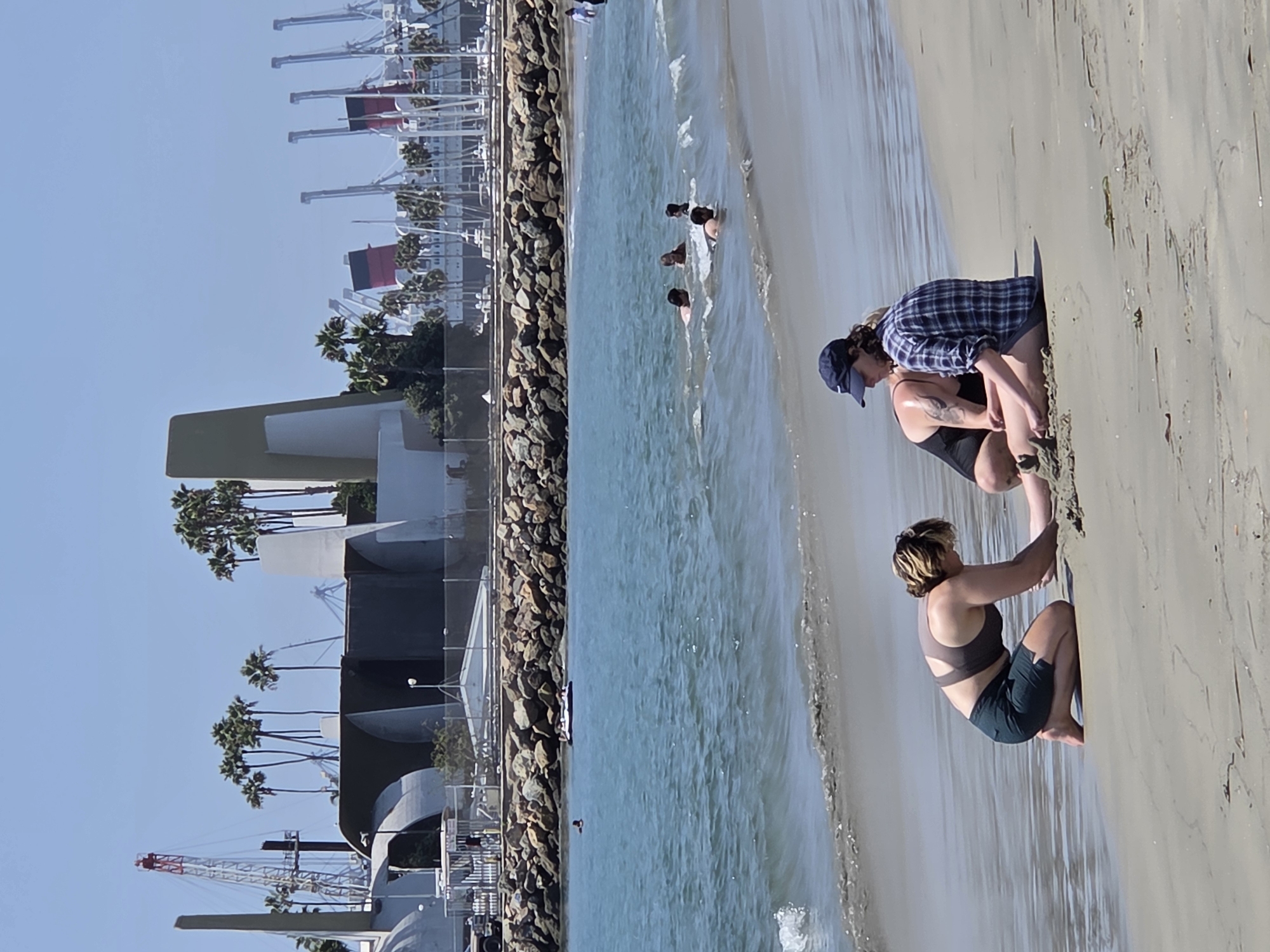 The image depicts a sunny day at the beach in Long Beach, California, with the Long Beach Waterfront visible in the background. In the foreground, three individuals are on the sand. Two women are kneeling and interacting with the sand, with one wearing a dark sports bra and green shorts, and the other wearing a black swimsuit. A third person, wearing a blue baseball cap and a plaid shirt, is also kneeling near them. The ocean is in the middle ground, with several people swimming and a small inflatable raft in the water. The background features the Long Beach Waterfront, characterized by its distinctive architecture, palm trees, cranes, and docked boats and ships, including the iconic Queen Mary with its red funnels. The water is blue and calm with gentle waves lapping at the shore.