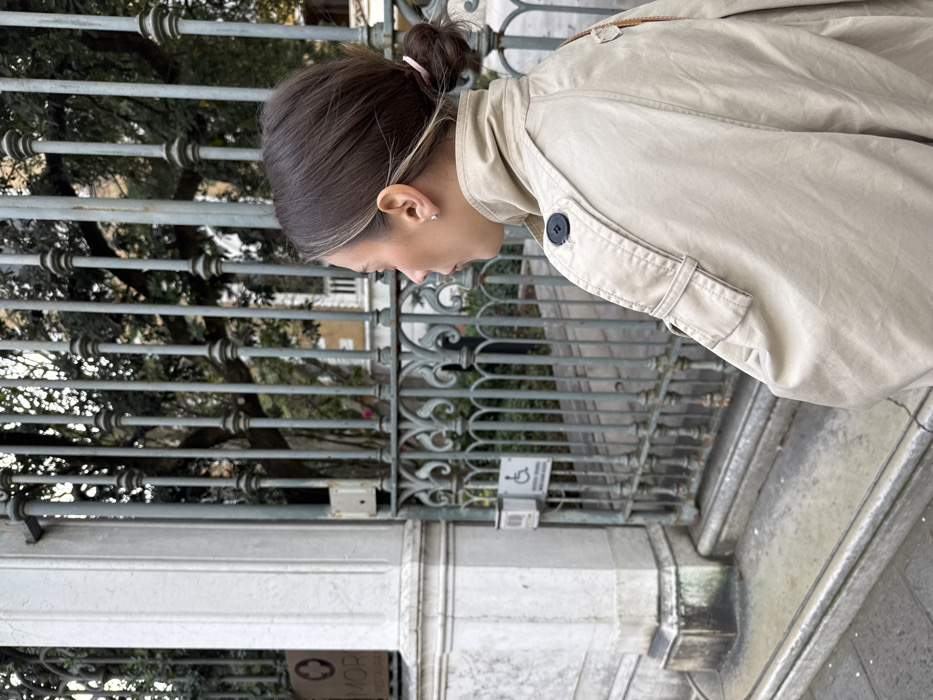 A female subject is captured in profile, facing left, standing on a stone pathway in front of a closed, ornate grey metal gate. She wears a light beige trench coat with a raised collar, a visible dark button on a shoulder epaulet, and her medium brown hair is styled in a low bun secured with a light pink elastic. A small stud earring is visible in her right ear. To the immediate left, a weathered white stone pillar features a vertical sign with a red cross logo above the text "MOP" and "RESTAURANT." On the adjacent gate, a white sign displays a wheelchair accessibility symbol and partially legible text. Beyond the gate, a dense garden with green foliage and indistinct building elements are visible. This location is identified as the entrance to the Hotel Metropole Venezia on Riva degli Schiavoni, Castello, Venice, Italy.
