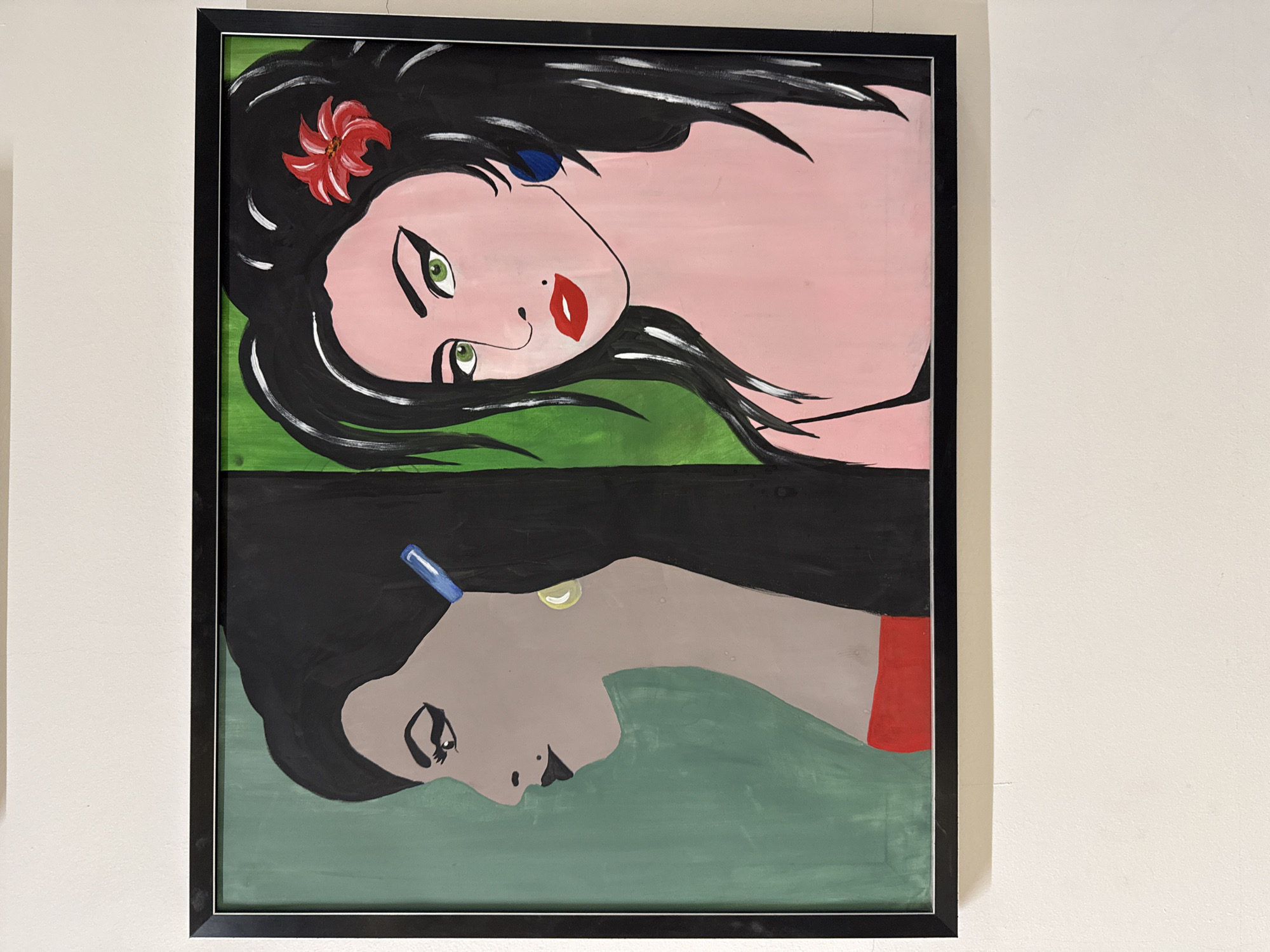 A framed painting hangs on a white wall. The painting is divided into two horizontal sections, with a portrait in each section. The upper portrait is of a woman with fair skin, black hair adorned with a red flower and a blue earring, green eyes, and red lips. She appears to be reclining on green fabric. The lower portrait is of a woman with grey skin, black hair with a blue barrette, and a gold earring, also reclining on green fabric. The painting is encased in a black frame. The location is a white-walled interior space, likely in a building in Izhevsk, Russia, based on the user query.