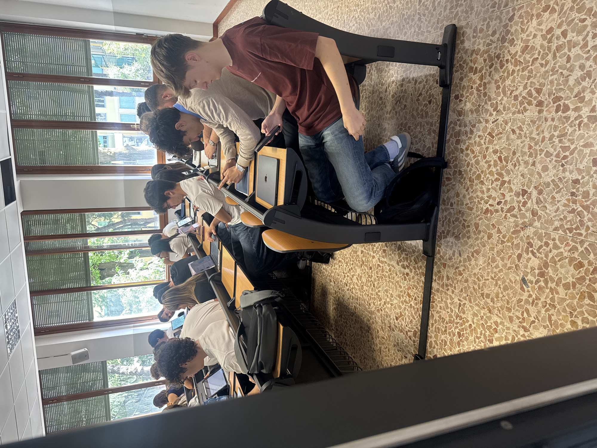The image captures a classroom or lecture hall from a slightly elevated perspective, looking down a row of students. Numerous young adults, primarily male, are seated at individual desks with attached chairs, engaged in an activity involving digital devices.
The students are focused on laptops or tablets, which are open on their light wooden desks. Several Apple MacBooks are clearly visible, identifiable by their iconic logo. One student in the mid-ground, wearing a maroon t-shirt and jeans, is standing or kneeling on his chair, leaning over his desk and looking intently at a device. Another student nearby is pointing at a screen, suggesting discussion or collaboration. Backpacks are stowed beneath some of the chairs.
The setting is bright, with ample natural light entering through a wall of large windows. These windows feature exterior slatted blinds, common in Italy. Beyond the windows, green trees and parts of distant buildings are visible, indicating an outdoor urban or campus environment. A white speaker is mounted on the wall between window sections. The floor is a distinctive terrazzo tile with a speckled pattern of light brown, beige, and white. The ceiling has rectangular light fixtures. The scene depicts a daytime academic or training session in Milan, Italy. No specific legible text beyond the Apple logo is visible.