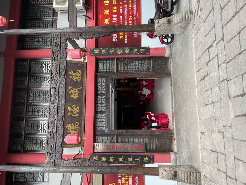 The image shows a red brick building in Chongqing, China at latitude 29.699, longitude 106.789, with an ornate wooden doorway that has a red lantern hanging from the top. The doorway is flanked by two large stone pillars, and there is a sign above the doorway that reads "Hong Yun Restaurant." The interior of the restaurant is visible through the doorway, and there are several tables and chairs set up. A single person, wearing a black jacket, can be seen sitting in a red chair at a table in the restaurant. Outside the doorway, a motorcycle is parked on the cobblestone street. There are advertisements on the wall in both Chinese and English, including information about the restaurant's capacity and contact information. There is a second red lantern hanging above the street side of the sign. A large red lantern hangs from a wooden pole outside the front of the building. The building is decorated with black painted wooden frames surrounding windows and doors.