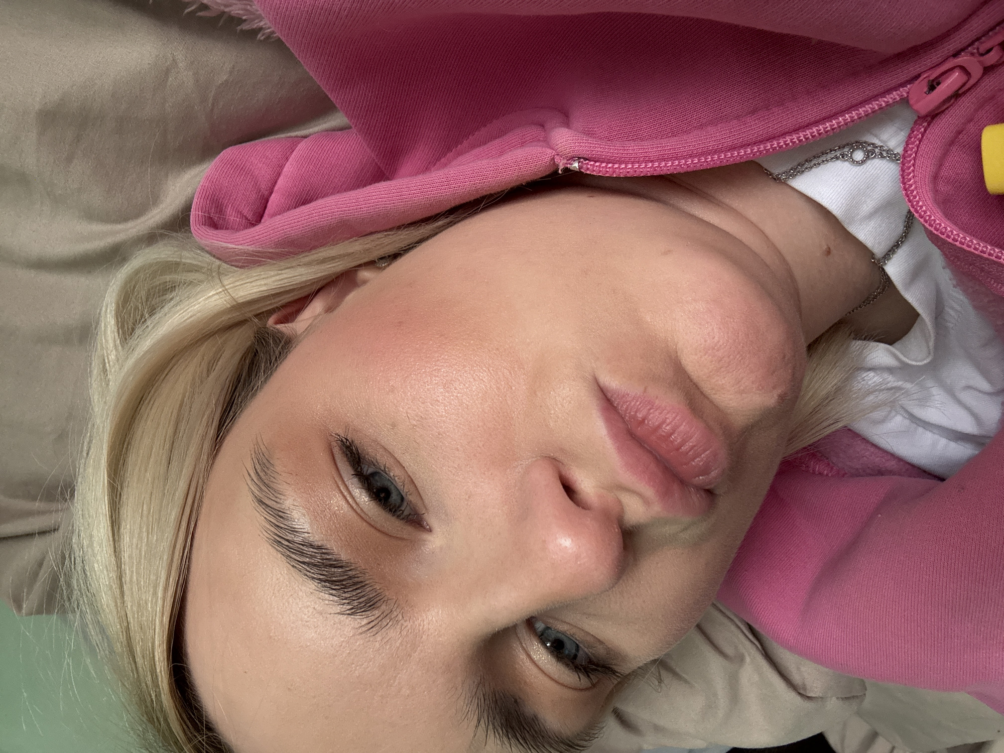 A close-up, angled selfie shows a fair-skinned person with light blonde hair and blue eyes. The person is wearing a pink hooded sweatshirt, and a white shirt is visible underneath, along with a silver necklace. The background appears to be beige fabric. The location is Unknown, Unknown.