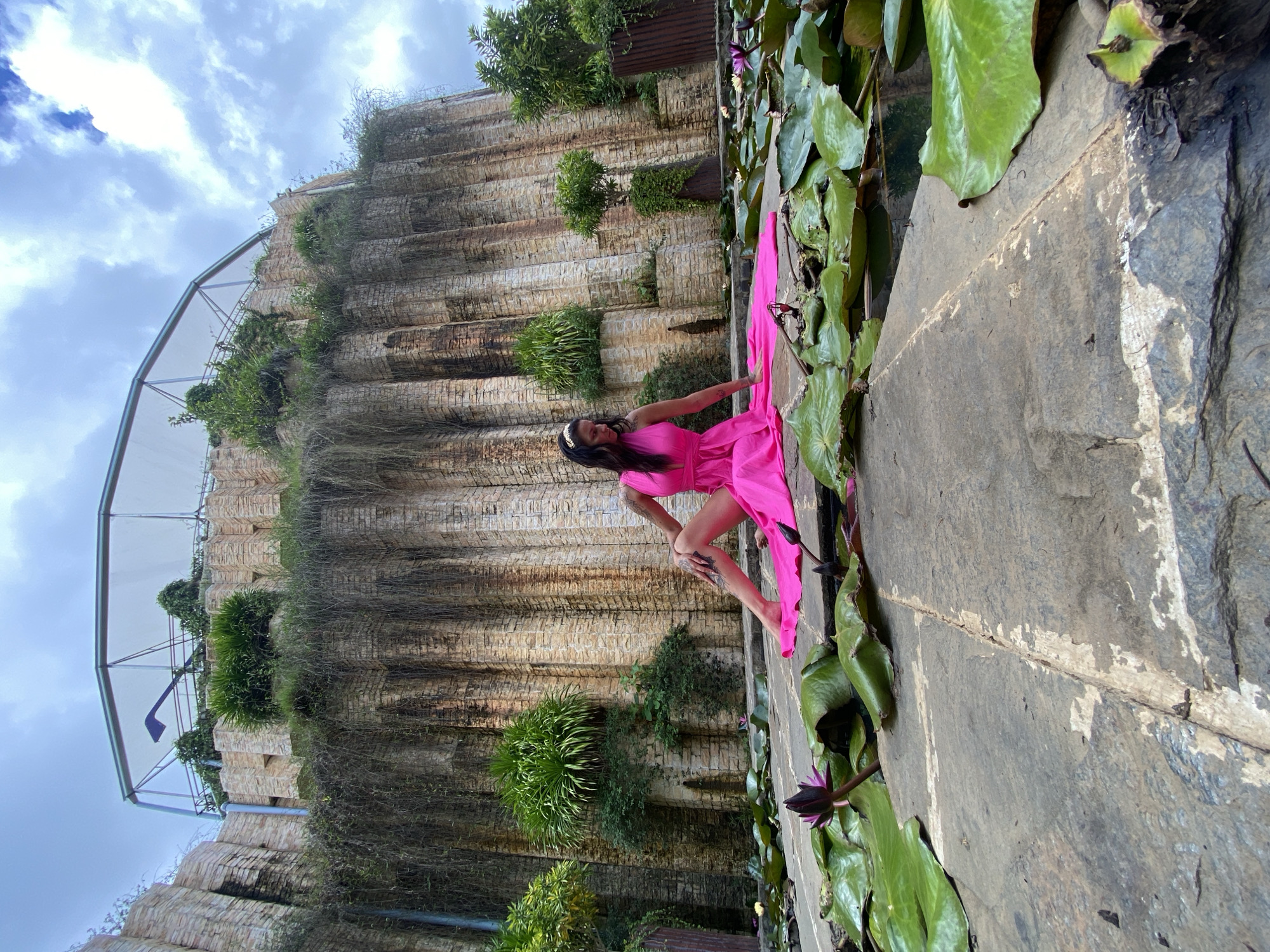 In Cam Ly - Đà Lạt, Vietnam, a woman in a vibrant pink dress sits gracefully on large, flat stones surrounding a lily pond. Her dress, with a flowing train, drapes across the stones and into the water, where large green lily pads and a few dark purple lotus buds are visible. Behind her, a towering, textured wall constructed of rough-hewn stones rises, draped with cascading vines and tufts of greenery. The upper portion of this structure features a netted canopy supported by metal beams, suggesting an outdoor recreational area or a unique architectural element. The sky above is a mix of bright blue and dramatic, cloudy formations.
