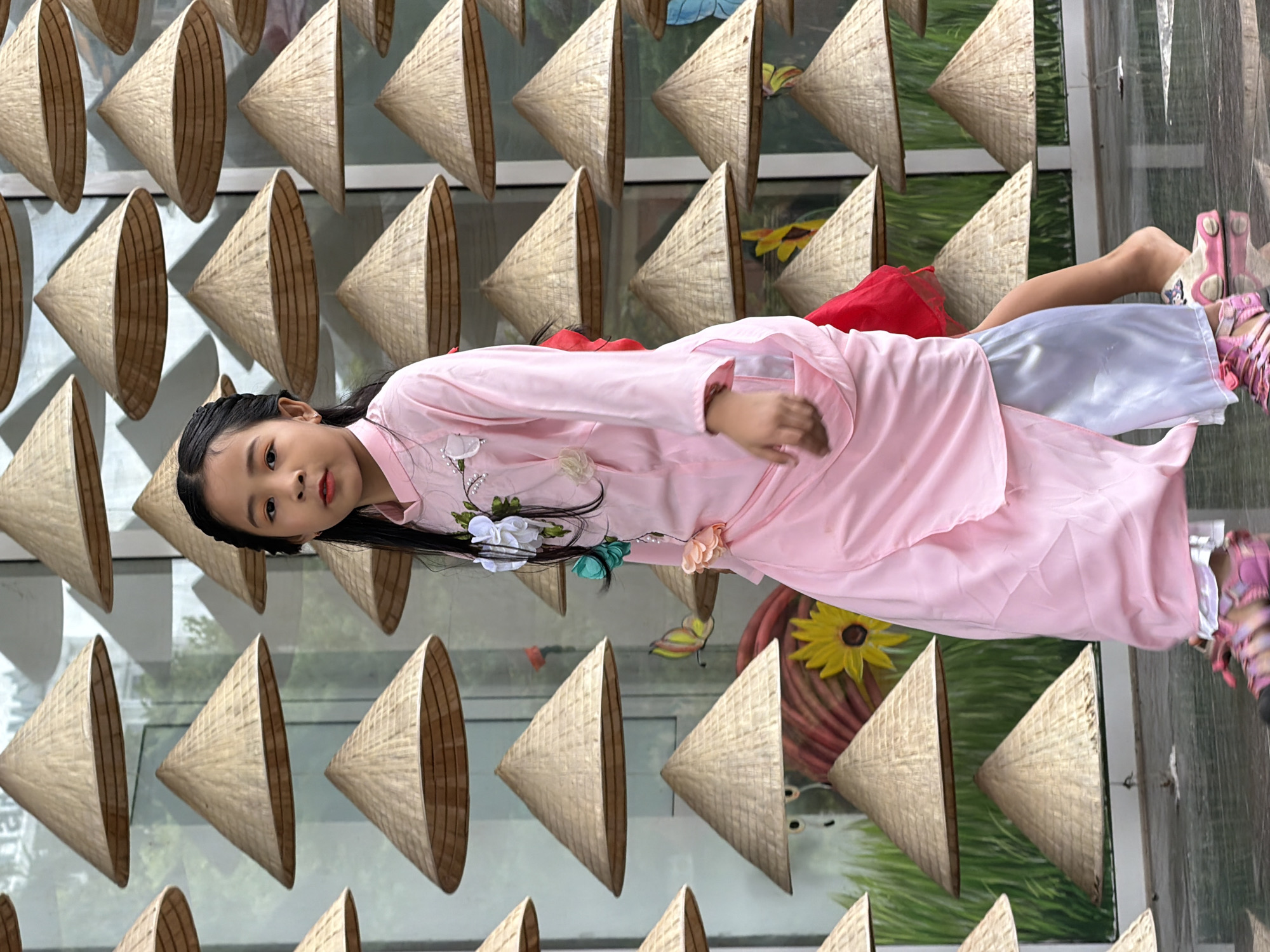 A young girl with dark hair, wearing a pink dress and colorful sandals, walks past a display of conical hats. The hats are arranged in rows against a backdrop that appears to be a wall or large window with a mural of grass and a yellow flower. The girl is looking towards the camera. The scene suggests an outdoor event or cultural display in a city, possibly in Vietnam.
