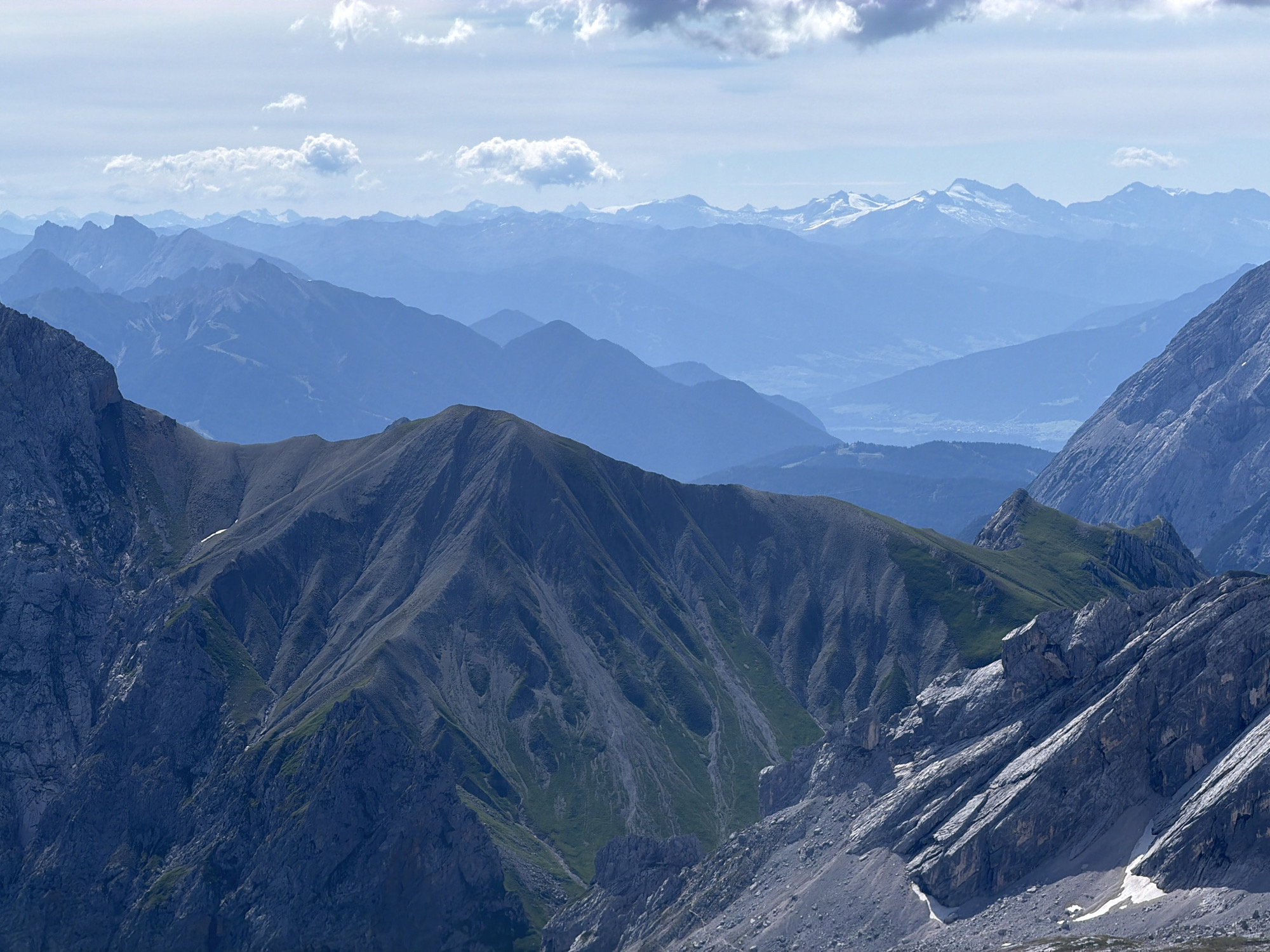 This is an outdoor, wide-angle, daytime, scenic photograph of a mountain range. The image captures multiple layers of mountains receding into the distance, with a clear sky featuring scattered white clouds overhead. The foreground features rugged, rocky slopes with patches of green vegetation, characteristic of a high alpine environment. The middle ground displays a prominent, dark-colored mountain with steep, textured slopes and visible gullies, also interspersed with green grass. Further back, numerous mountain ranges appear in varying shades of blue and gray due to atmospheric perspective, with the furthest peaks possibly capped with snow or ice. The overall impression is one of vastness and natural grandeur. The scene is devoid of any people, structures, or signs of human activity, presenting a purely natural landscape. The lighting suggests a bright, sunny day, casting shadows that highlight the topography of the mountains. This image likely depicts a view from a high vantage point within the Alps. While Garmisch-Partenkirchen is a town situated in the Bavarian Alps and is known for its mountainous surroundings, the specific location within or near Garmisch-Partenkirchen cannot be determined solely from the visual information in this photograph.