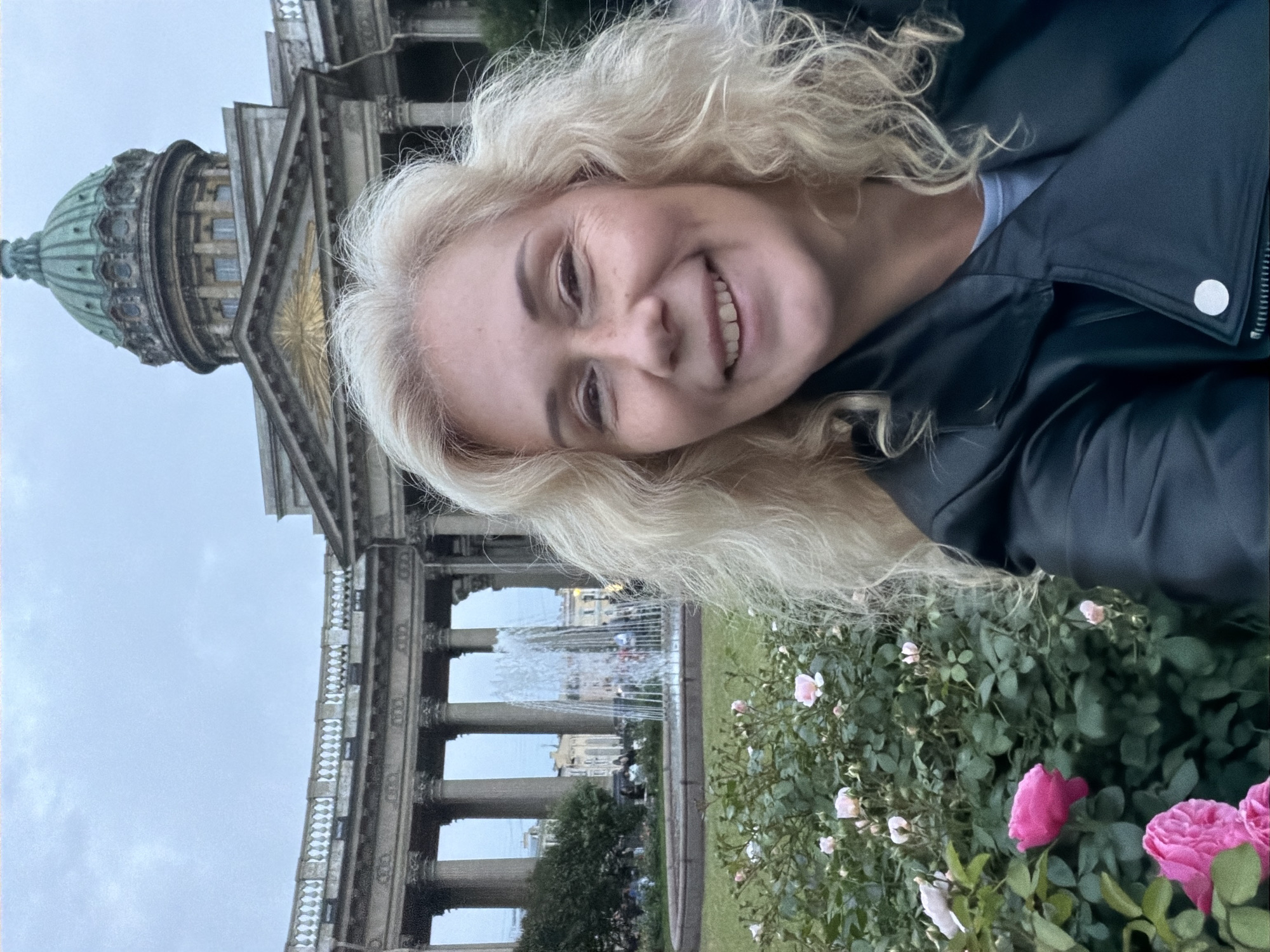 A candid selfie is captured in front of the Kazan Cathedral in St. Petersburg, Russia, on an overcast day. The woman in the foreground has blonde, curly hair and is wearing a black leather jacket over a light-colored shirt. She is smiling broadly, her eyes looking at the camera. In the middle ground, a large, ornate fountain sprays water upwards, surrounded by a circular pool. Behind the fountain, the impressive facade of the Kazan Cathedral features a grand colonnade and a prominent green dome. The foreground also shows a bush with pink roses, some in bloom. People can be seen in the background, dispersed around the fountain and the cathedral's grounds.