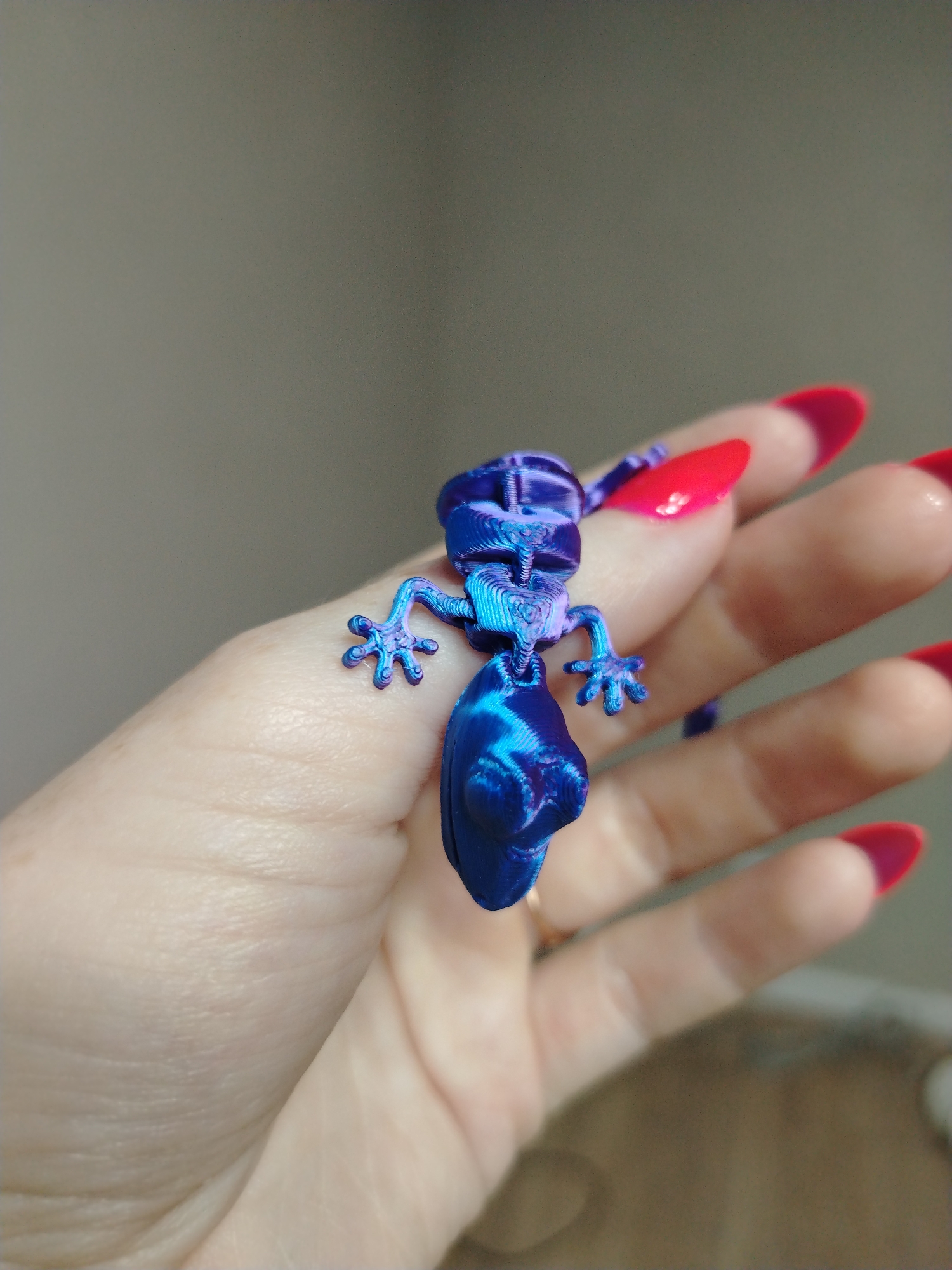 A person's hand is holding a small, blue-and-purple, articulated lizard figure. The lizard is positioned head-up, with its limbs spread against the palm. The person has long, manicured nails with a red-and-pink gradient. The background is a neutral, light gray-brown wall.
There is no information in the image to indicate this scene is located in Izhevsk, Russia.