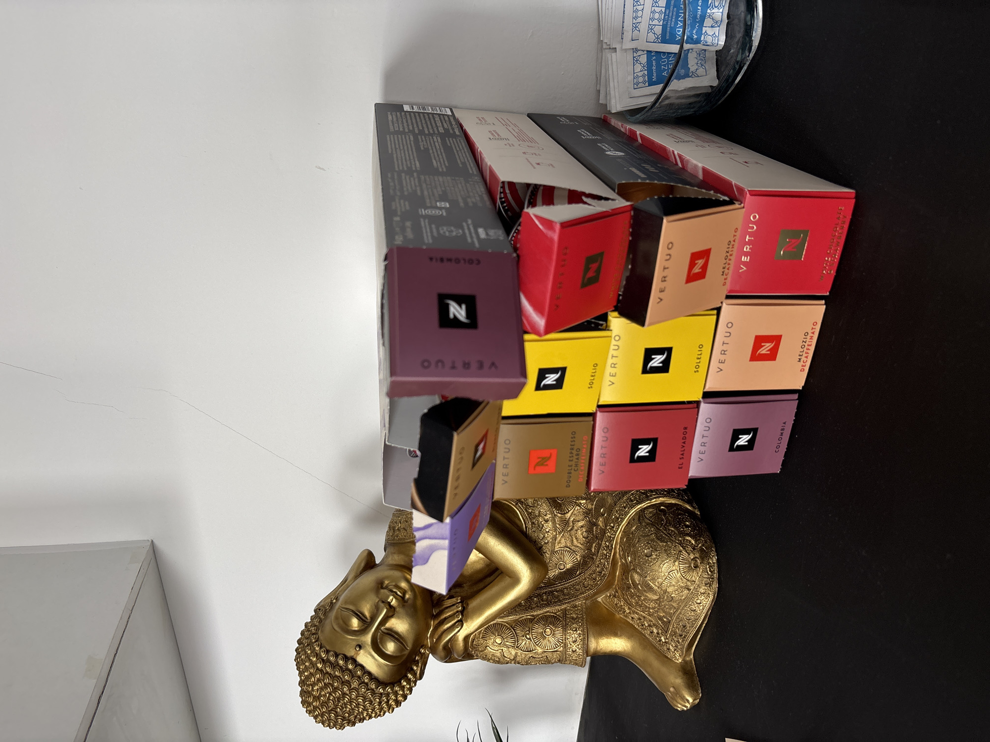 This indoor scene, located in the city of Cholula, Mexico, displays a golden Buddha statue on a black surface, positioned to the left. The Buddha is depicted reclining with its head resting on its right arm, eyes closed, and adorned with ornate golden robes. To the right of the statue, a vertical stack of Nespresso Vertuo coffee capsule boxes is arranged, featuring various colors such as purple, yellow, red, orange, and grey, with several boxes partially open. Labels visible on the boxes include "Double Espresso Chiaro," "Solelio," "El Salvador," "Colombia," "Melozio Decaffeinato," and "White Chocolate & Strawberry." To the far right, a clear glass bowl contains numerous small white packets, some labeled "Member's N Azúcar Fina" (fine sugar) and others "Edulcorante" (sweetener). The background consists of a plain white wall, which shows a faint, thin vertical crack in the upper right quadrant. A light-colored shelf or ledge is visible in the top left corner of the frame.