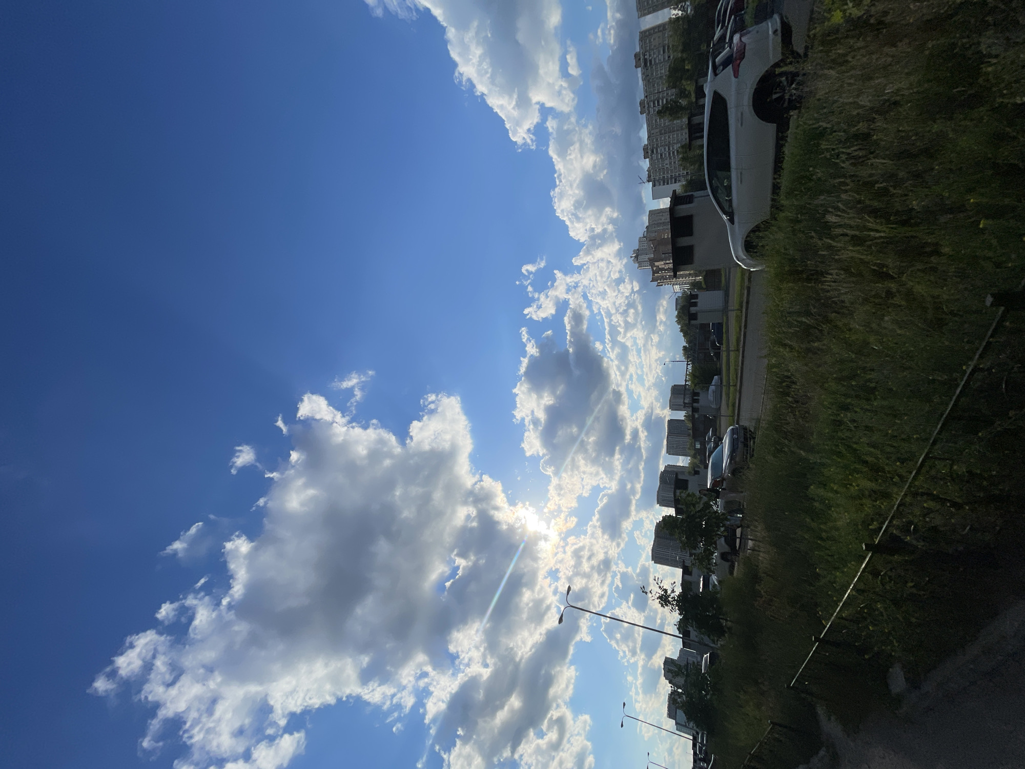 This is a vertical, low-angle photograph of a sky filled with cumulus clouds and a bright sun peeking through them. Below the sky, the scene depicts a city landscape. In the foreground, there is a patch of tall, dry grass bordering a dark metal fence. To the right of the grass is a paved area with a white SUV parked. Further back, a road is visible with several cars, including a silver sedan. Beyond the road, a row of tall, residential buildings, typical of Kyiv's urban architecture, stretches across the horizon. Streetlights are also visible. The sun's rays are creating a lens flare effect, adding a bright glow to the sky. The overall scene captures a sunny day in a residential area of Kyiv, Ukraine, possibly in the late afternoon or early evening given the angle of the sun.