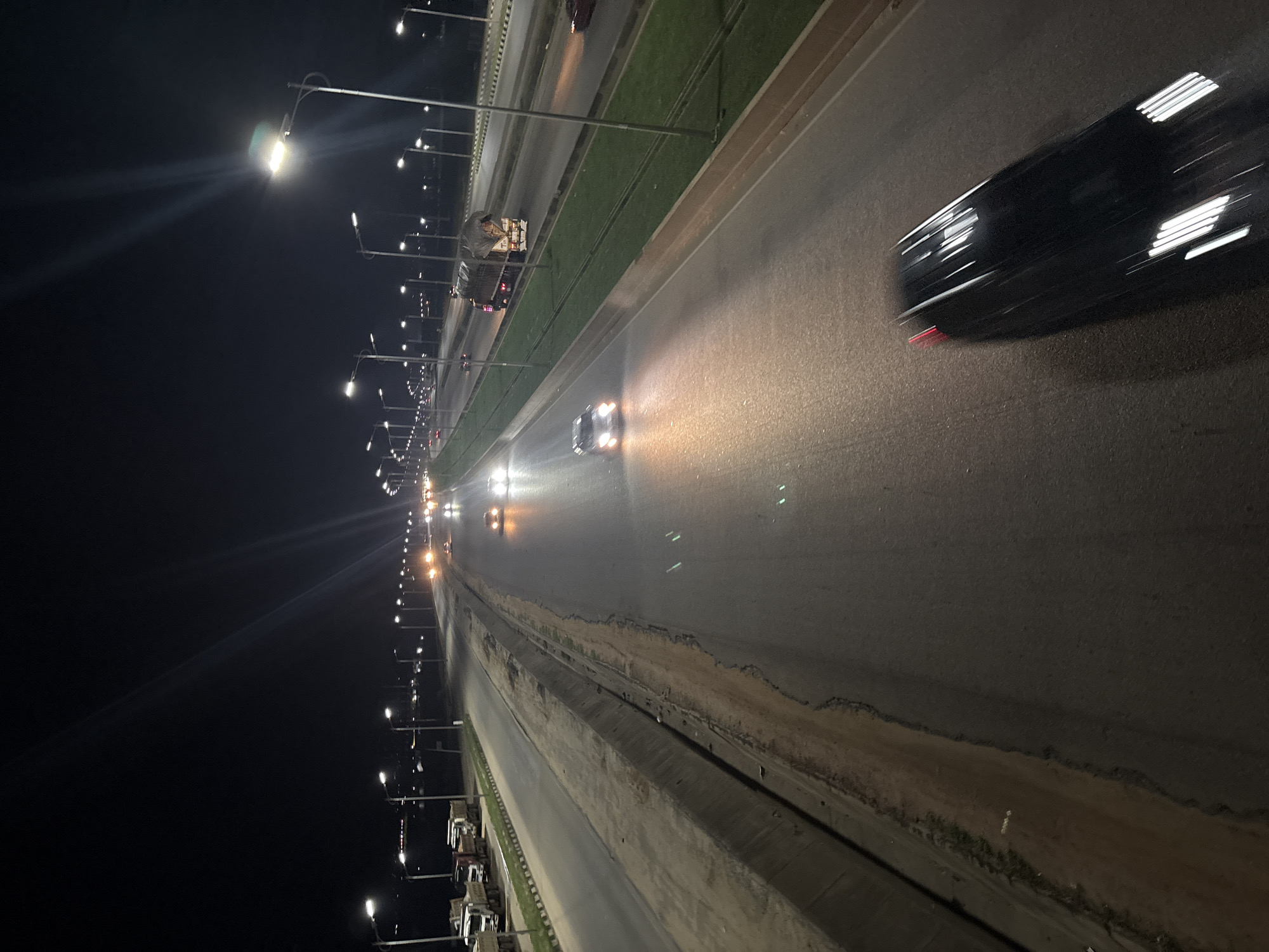 The image depicts a highway at night in Angwan Sayawa, Nigeria, with multiple lanes of traffic illuminated by streetlights. Several vehicles, including cars and trucks, are visible on the road. Some cars have their headlights on, and a few appear to be in motion, indicated by motion blur. The environment consists of a paved roadway with a grassy median and concrete barriers. Streetlights line the highway, casting light on the road and surrounding area. The scene suggests a typical night on a busy urban or suburban highway.