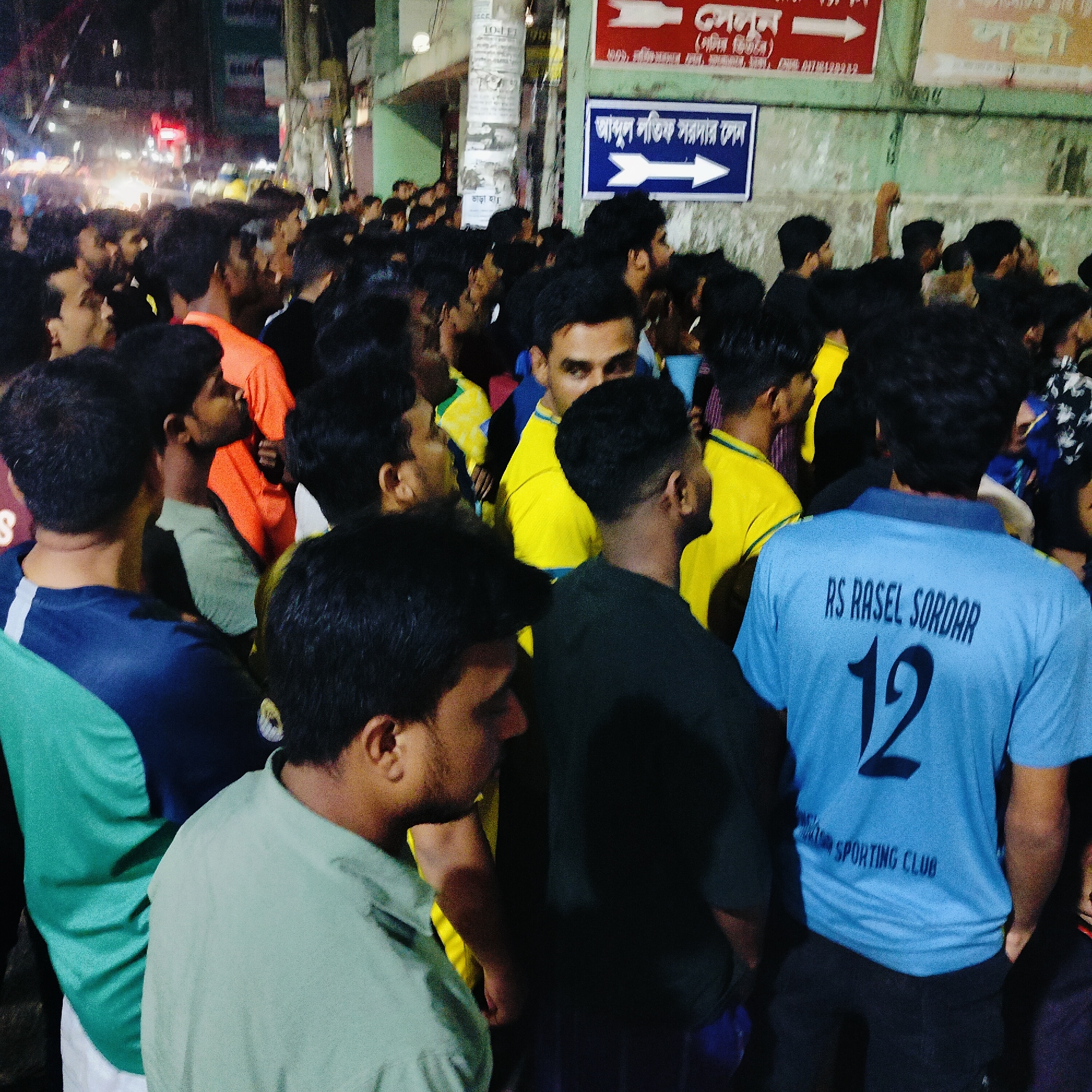 The image depicts a crowded street scene at night in Dhaka, Bangladesh. A dense gathering of men, primarily young adults, are visible in the foreground and background, suggesting a public event or gathering. The lighting is dim and artificial, with streetlights and building lights illuminating the area. Several individuals are wearing t-shirts with visible text and numbers. Specifically, one person in the foreground wears a light blue t-shirt with the text "RS RASEL SORDAR" and the number "12" on the back, along with "SPORTING CLUB" below. Other individuals are wearing brightly colored t-shirts, including yellow and orange. In the background, a blue and white sign with Bengali script and an arrow pointing to the right is affixed to a wall, indicating directions or information. The environment appears to be an urban street, with buildings and posters visible along the side of the road. The overall impression is of a lively, densely populated urban environment at night.