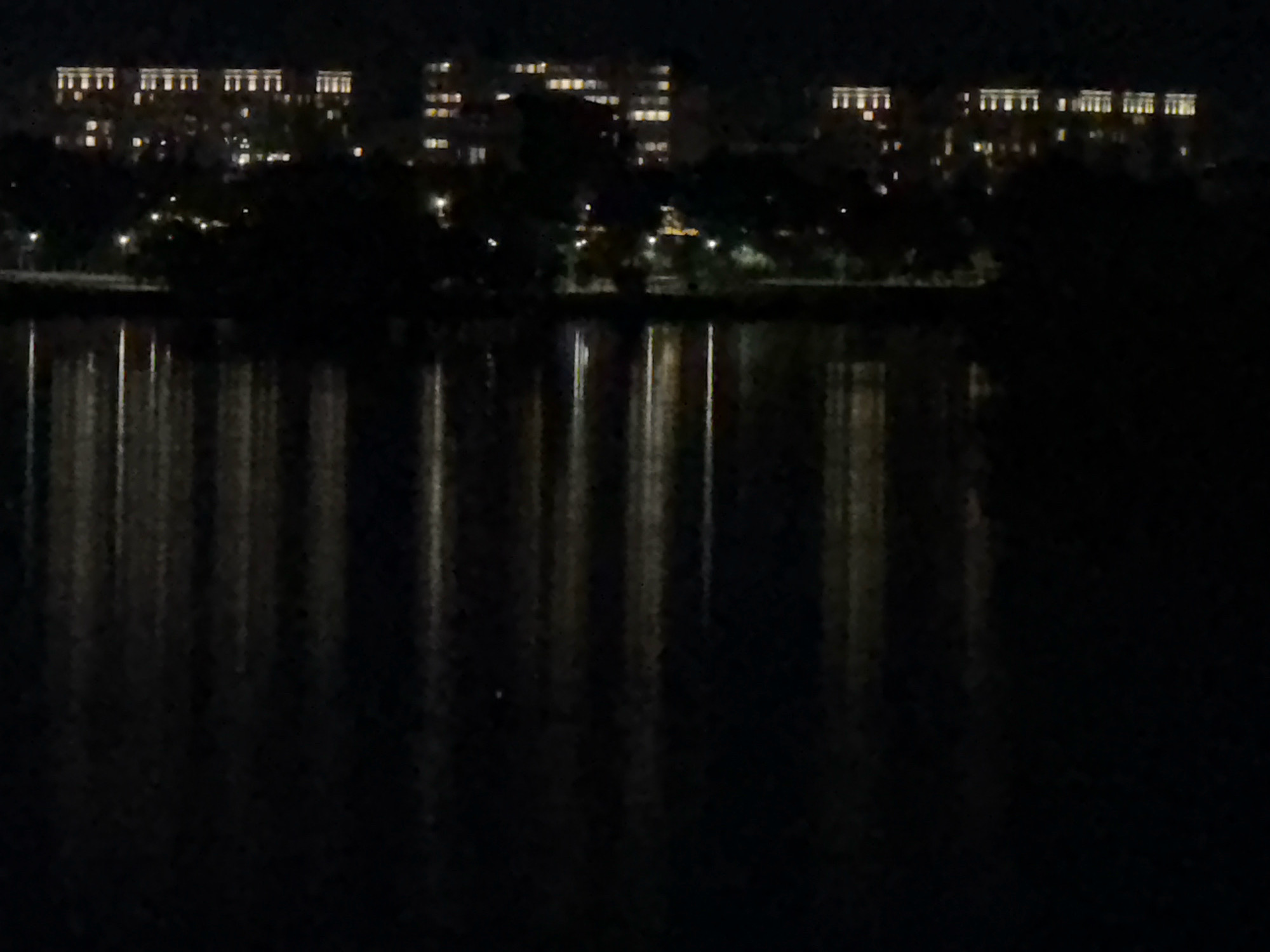 The image depicts a dark, nighttime scene of buildings and their reflections in water. Several multi-story buildings with numerous illuminated windows are visible in the upper portion of the image. These buildings appear to be situated along a waterfront, as their lights are reflected in the dark water in the foreground. Trees and foliage are visible between the buildings and the water, obscuring some of the lower parts of the structures. The overall impression is that of an urban or suburban environment at night, with artificial lights from buildings contrasting with the darkness of the natural surroundings. The specific location in Pavlodar, Kazakhstan, is not discernible from the visual information provided in the image alone.