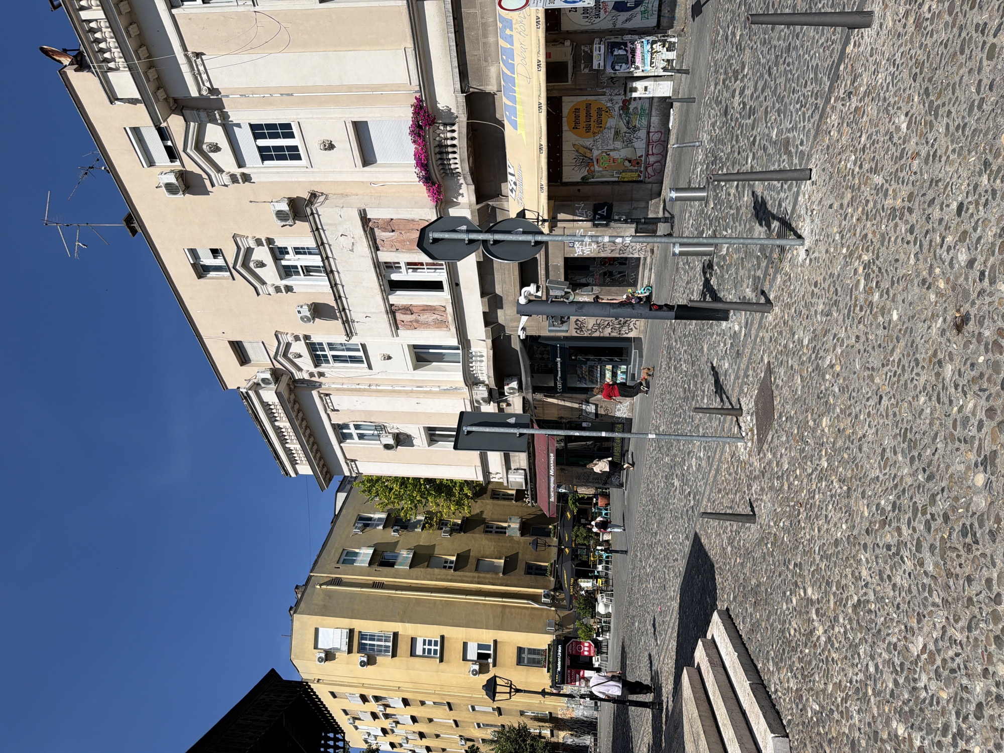 This is a sunny, outdoor, street-level view of a cobblestone plaza in Belgrade, Serbia. The plaza is flanked by buildings, with a clear blue sky overhead.
On the left side of the image, a light yellow building stands tall. Its facade features numerous windows and balconies, and air conditioning units are visible on its exterior. In front of this building, there are steps leading up to a walkway, a lamppost, and a Western Union sign. A lone man in a white shirt and dark pants walks away from the camera on the cobblestones.
To the right of the yellow building, a cream-colored building with ornate architectural details, including classical moldings and a balcony with pink flowers, dominates the scene. The ground floor of this building houses shops with awnings. One awning is yellow and displays the word "AMANI." Various posters and graffiti adorn the storefronts. Several people are visible walking in front of these shops, including a woman in a red shirt and a person walking a small dog. A set of traffic lights are visible, and a traffic sign with two dark grey discs is placed on a pole in the plaza.
Further into the plaza, tables and chairs are set up, suggesting an outdoor seating area for cafes or restaurants. The cobblestone pavement is the dominant ground covering. The scene appears to be a bustling urban environment on a clear day.