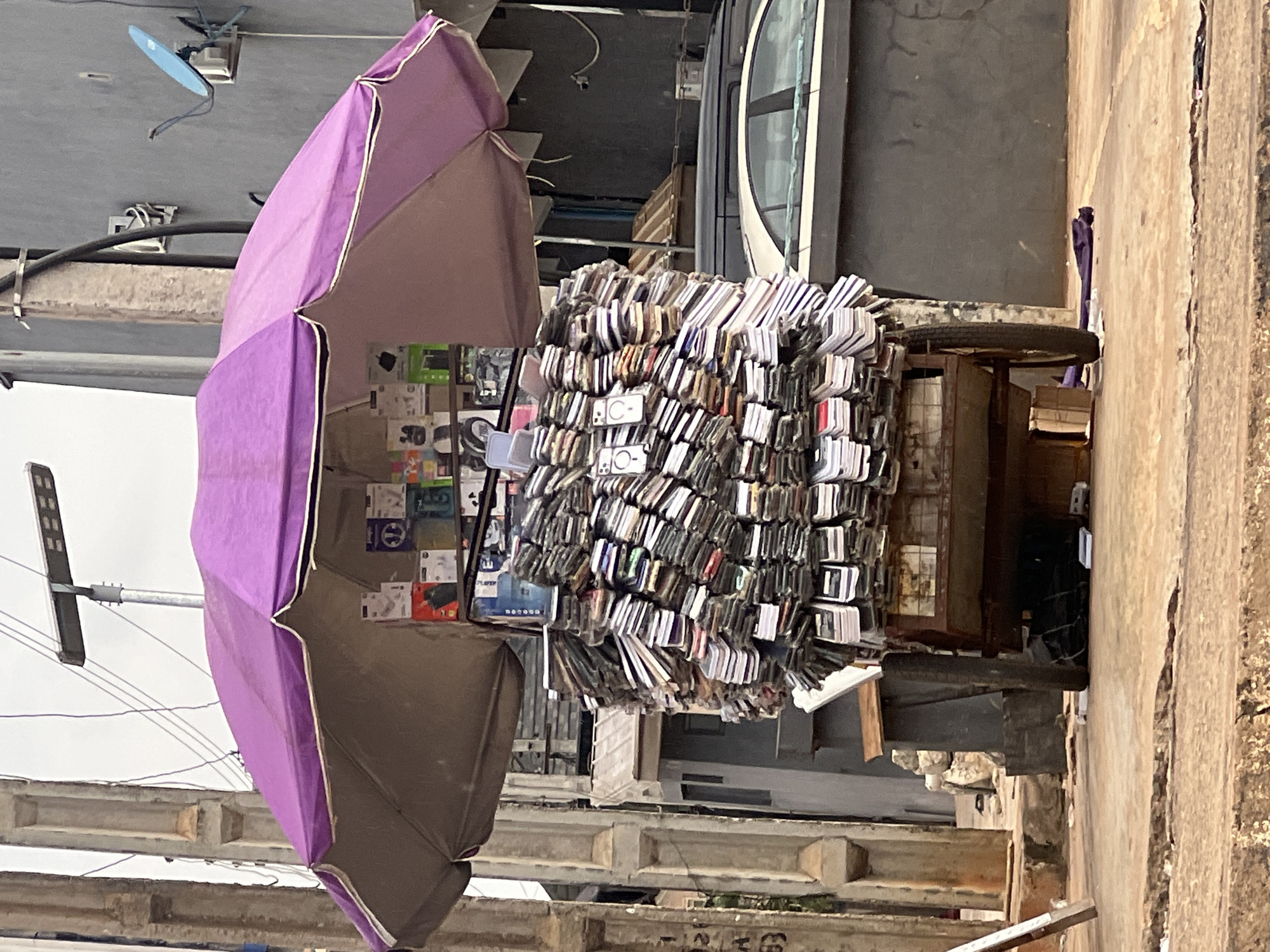 In Okpanam, Nigeria, a mobile vendor operates a cart laden with a vast quantity of phone accessories, primarily phone cases, under a large purple umbrella. The cart, equipped with two wheels, is positioned on a paved street, likely a roadside stall. The accessories are meticulously stacked, forming a towering display of various colors and designs. Above the phone cases, several boxes of smaller electronics, possibly chargers or earphones, are visible, also displayed under the umbrella. The background reveals a typical urban environment with buildings, utility poles, electrical wires, and a satellite dish. A dark-colored car is parked nearby, and the ground is a mix of concrete and dirt. The scene captures a common street vending setup in Nigeria, showcasing a dense collection of mobile phone accessories.