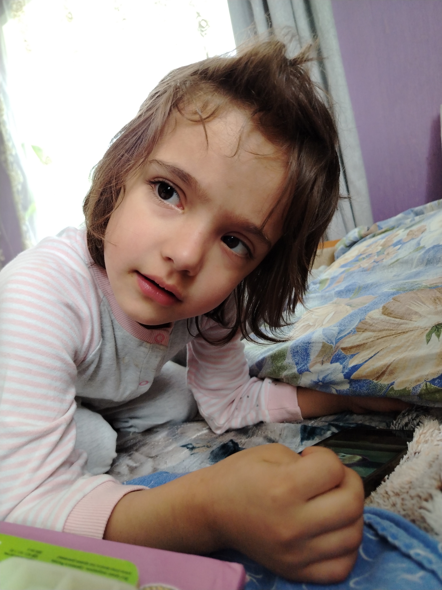 A young girl with brown hair, wearing pink and grey striped pajamas, is kneeling on a bed covered with floral-patterned bedding. She is looking to the left with a slight frown. Her right hand is clenched into a fist, partially obscuring a black smartphone lying on the bed. In the background, there's a window with light filtering through and a purple wall. It is not possible to verify if this image was taken in the city of Гожан, Russia, with the available information.