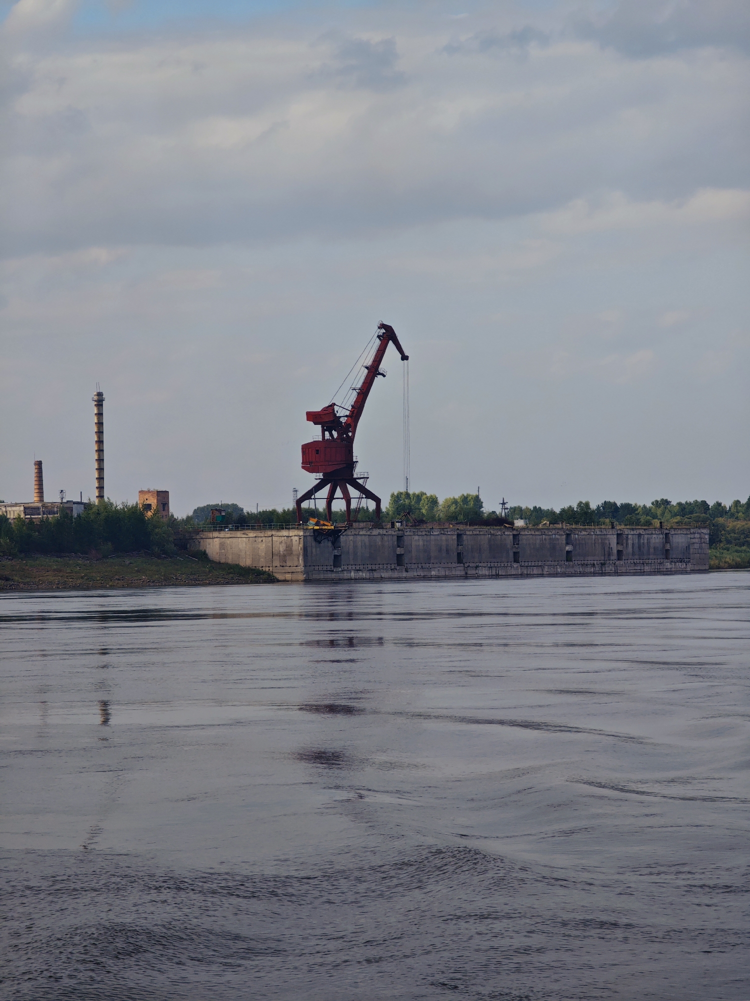 A wide, calm river flows in the foreground, reflecting the overcast sky and the industrial structures on the opposite bank. Dominating the center of the frame is a large, red gantry crane situated on a concrete dock. Behind the crane, a row of trees partially obscures a long, concrete structure, possibly part of a shipyard or port facility. To the left of the crane, on the distant bank, are several industrial buildings, including a tall, cylindrical smokestack and two smaller brick structures. The sky above is filled with soft, grey clouds, suggesting a cooler climate. The overall scene depicts an industrial waterfront with a prominent crane and port infrastructure. This appears to be a view from the river towards the industrial zone in Verkhnepashino, Russia, a port town located on the Yenisei River.