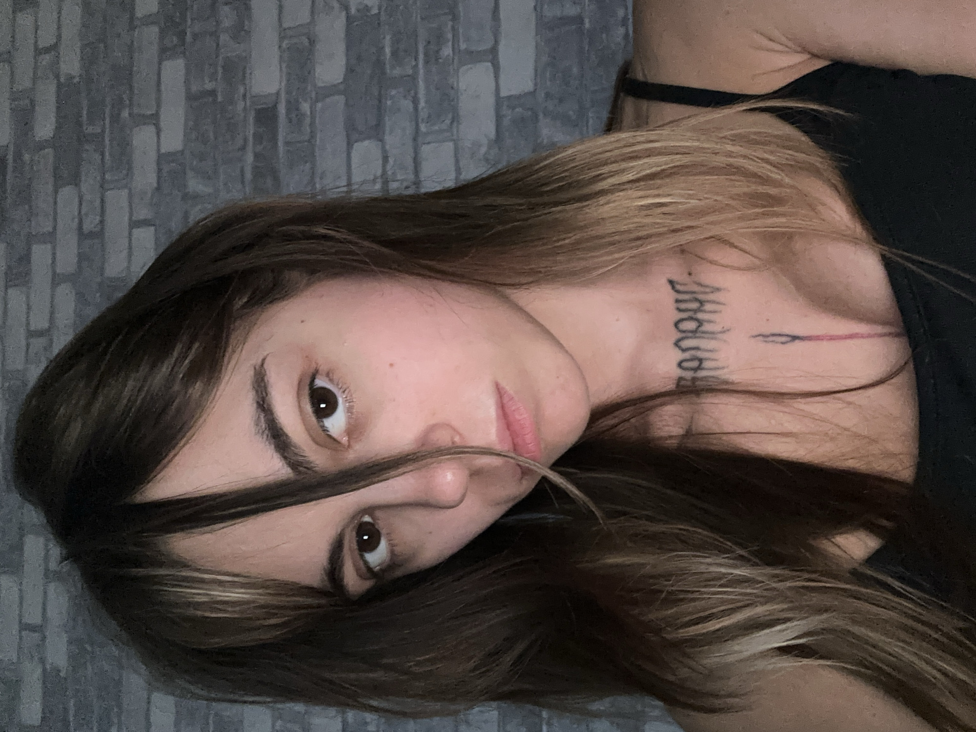 A woman with dark brown hair and bangs is captured in a close-up portrait. She is wearing a black tank top and has a tattoo on her chest and neck. The background consists of a gray brick wall. The image does not provide any information about the location in Moscow, Russia.