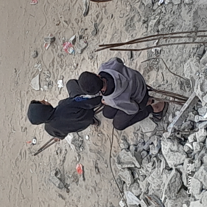 An overhead view captures two individuals seated amidst a debris-strewn landscape in Khan Yunis, Palestinian Territory. The ground is a mix of light-colored soil, sand, and abundant rubble, primarily fragmented concrete and rocks. The individual in the foreground, facing away from the camera, wears a light gray hooded sweatshirt, dark pants, and dark flip-flops, crouching on exposed, bent metal rods. Behind and to their left, another individual, wearing a dark hooded sweatshirt and dark pants, holds a long, dark, two-pronged gripping tool. This individual is also seated or crouched on similar metal rods. Both individuals are oriented towards the upper right portion of the frame. The immediate environment includes scattered refuse, such as fragments of red and white plastic wrappers, alongside numerous pieces of construction debris and protruding metal rebar. The background maintains a consistent appearance of disturbed earth and structural wreckage.