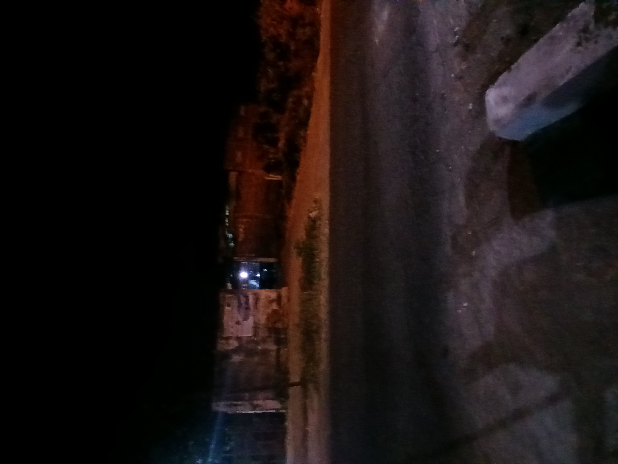 The image depicts a street scene at night in Kademangan Kulon, Indonesia. In the background, a dilapidated building with peeling posters is visible. Lights emanate from within the building, suggesting it is occupied or open. The foreground shows a paved road with a concrete barrier on the right side. The scene is dimly lit by artificial light sources, creating shadows and obscuring some details. The overall impression is one of a quiet, possibly neglected, urban environment at night.