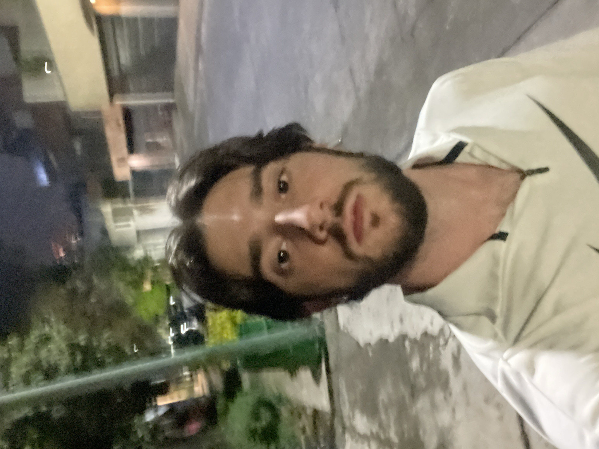 A selfie taken at night on a street in Huixquilucan de Degollado, Mexico shows a young man with dark hair and a beard looking directly at the camera. He is wearing a light-colored Nike jacket with a black collar and a large black Nike logo on the right side of his chest. The street appears to be paved and wet, reflecting the lights of the surrounding buildings and trees. The background shows houses and streetlights, indicating an urban or suburban environment. The overall lighting suggests it is nighttime, and the focus is on the man's face and upper body.