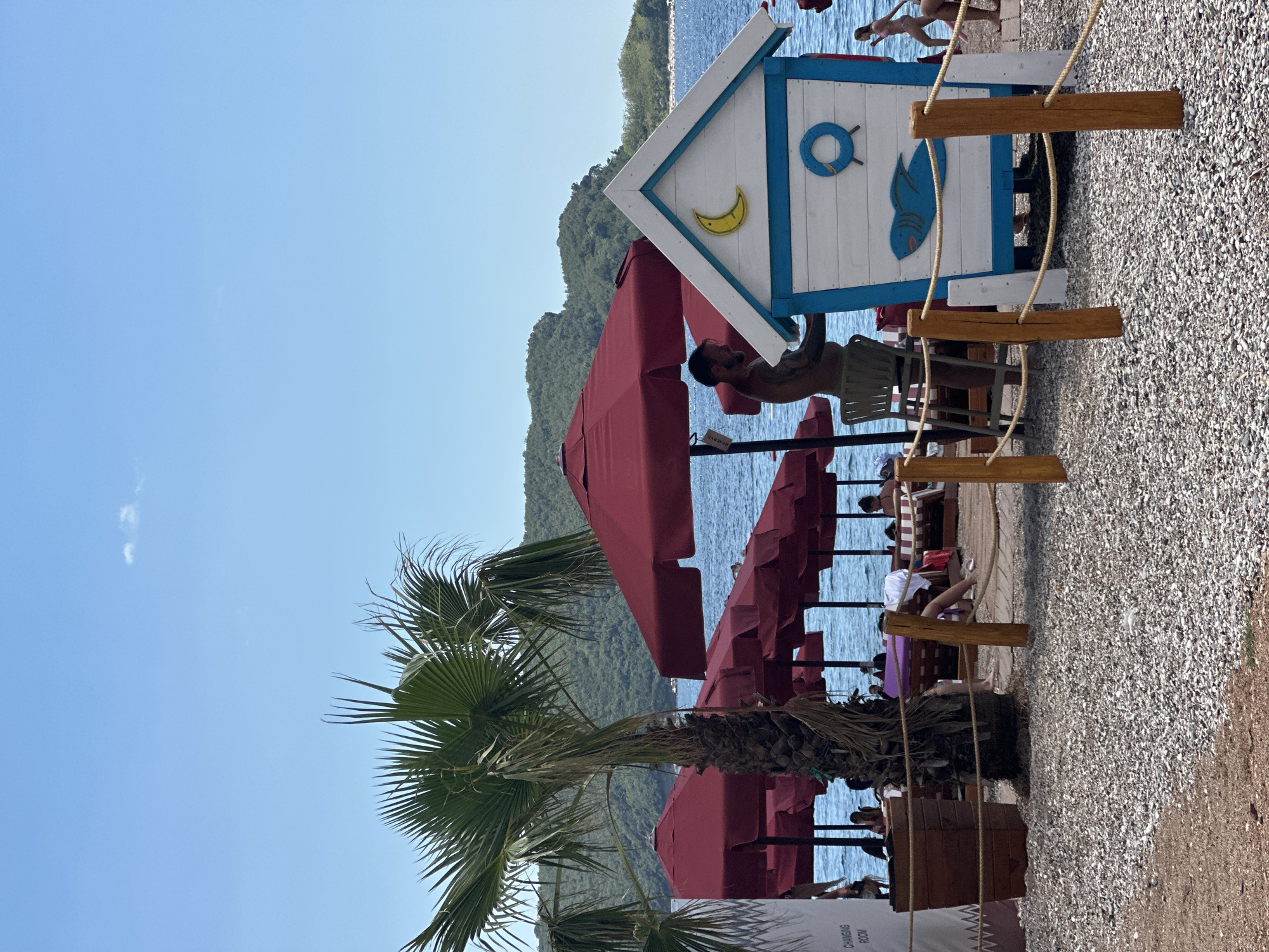 The image depicts a sunny day at a pebble beach in Budva, Montenegro. In the foreground, a small, decorative changing hut with blue and white trim, featuring a crescent moon and a fish motif, is positioned on the beach. Wooden posts and rope delineate a pathway. Behind the hut, a person stands near a chair, partially obscured by red beach umbrellas. Further back, more red umbrellas shade lounge chairs and beachgoers, with the blue Adriatic Sea and lush green hills in the background. A palm tree stands to the left of the scene. A sign indicating "CHANGING ROOM" is visible on the far left.