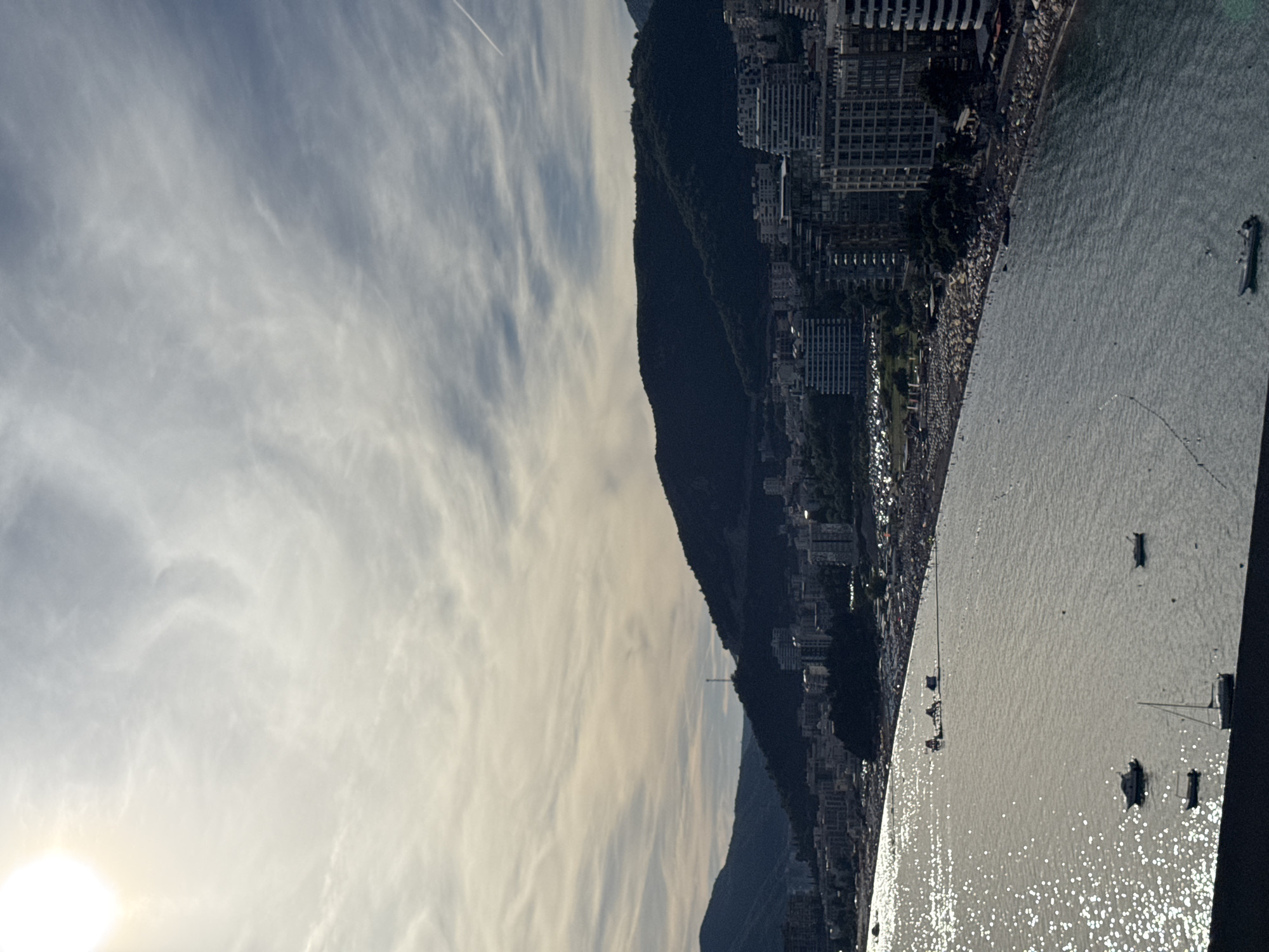 The image shows a wide, aerial view of the coastline in Rafailovići, Montenegro. The sun is visible in the upper left corner, casting a bright glare on the water. The sea is calm, with several boats and yachts anchored near the shore. The beach is lined with many people, and there are numerous buildings and hotels situated along the coastline, backing onto lush green hills. The sky is partly cloudy, with wispy cirrus clouds and a faint contrail from an aircraft. The scene depicts a bustling coastal resort town.