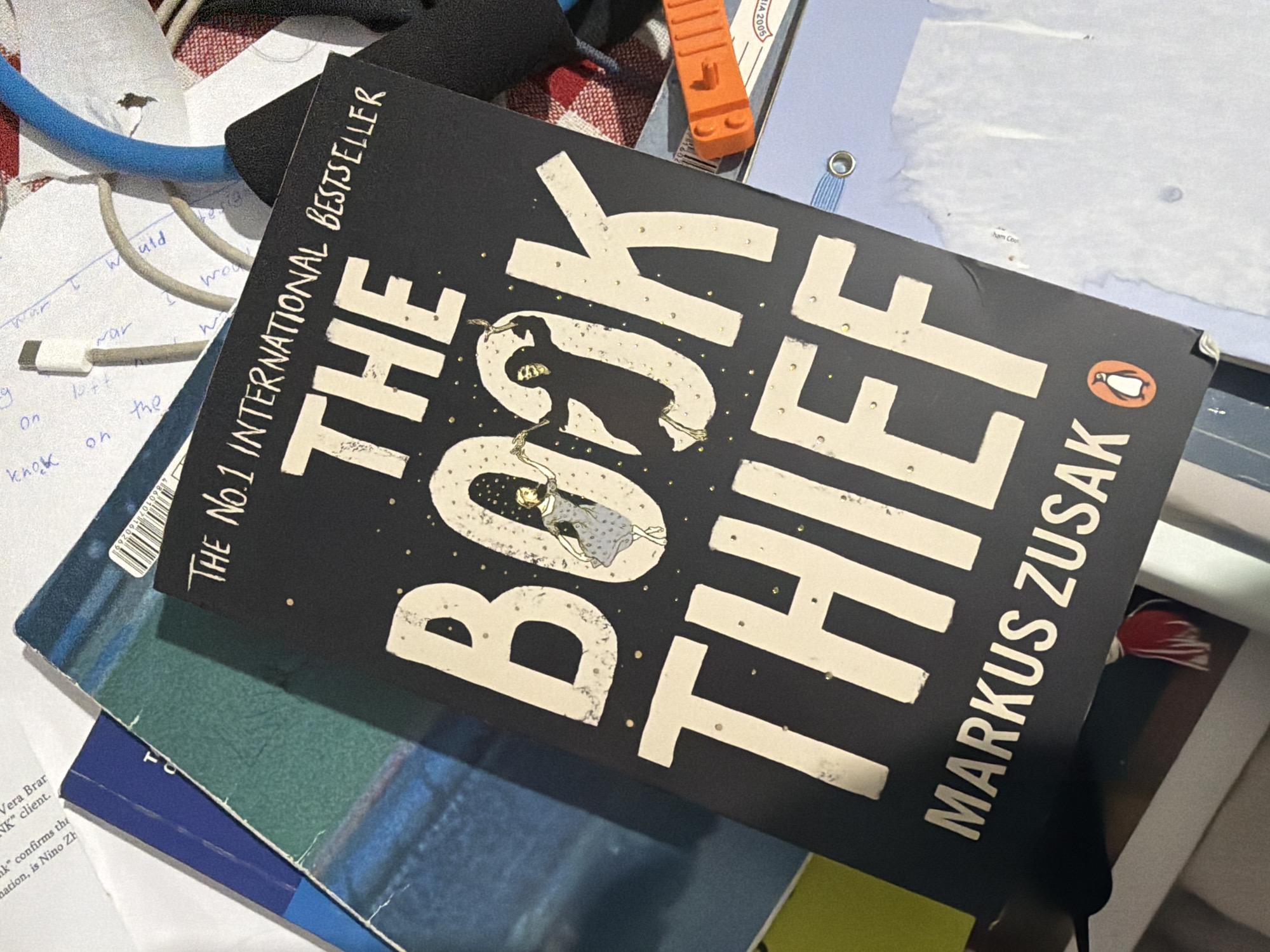 The image presents a close-up, top-down view of a cluttered surface, likely a desk or table. The central subject is a copy of "The Book Thief" by Markus Zusak. The book's dark cover features the title in large, cream-colored block letters. The word "BOOK" is uniquely designed, with an illustration of a small person in a patterned dress floating within its curves, surrounded by golden specks. The author's name, "MARKUS ZUSAK", is printed vertically along the right edge, and a small orange Penguin Books logo is visible. Above the main title, the text "THE NO.1 INTERNATIONAL BESTSELLER" is displayed.
Surrounding the book are various everyday objects. A blue cable and a white/grey cable are intertwined on the left, next to a crumpled white paper with handwritten notes like "would", "war", and "knock". In the upper right, an orange Lego brick and a light blue binder with a metal eyelet are visible. Below the main book, another book or document with a blue and dark green cover can be seen. On a white paper at the bottom left, printed text fragments such as "Vera Brar", "client", and "Nino Zh" are legible. A barcode with numbers "4 860010 107160269" is also partially visible. The setting suggests an indoor environment, possibly a personal study or work area, with no people present. The lighting is diffused, indicating either indoor artificial light or indirect natural light.