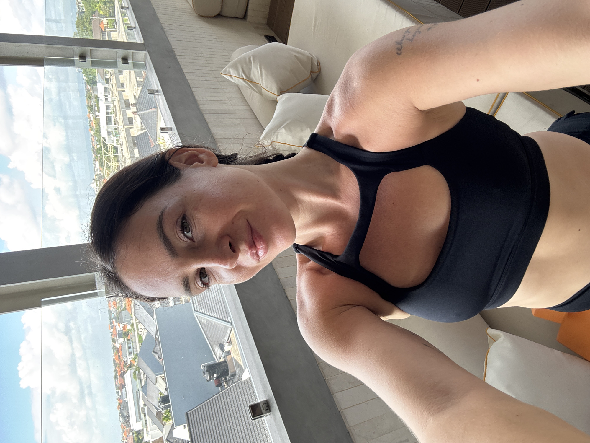 A woman with dark hair pulled into a braid and no makeup takes a selfie on a covered balcony overlooking a city. She is wearing a black sports bra and black shorts. The balcony has a gray railing and a glass barrier. Behind her, a beige couch with white pillows is visible. The city view shows rooftops, buildings, and a cloudy sky. The location is North Kuta, Indonesia.