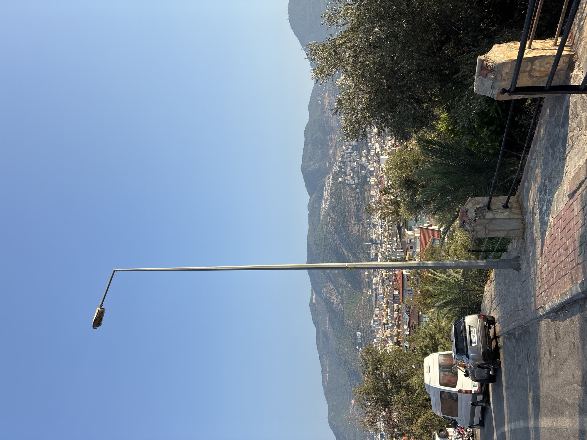 The image depicts a street scene overlooking the city of Alanya, Turkey, with mountains in the background. In the foreground, a silver SUV is parked on the right side of a paved road. To the left of the SUV, a white van is also parked. The road has a brick-patterned sidewalk on the right, bordered by a stone wall and a black metal railing. Beyond the railing, dense greenery and trees are visible. In the middle ground, the city of Alanya stretches out, with numerous buildings and rooftops, including many solar water heaters. The background is dominated by rolling green mountains under a clear blue sky. A tall, gray street lamp stands prominently in the center of the frame. The sun appears to be low, casting long shadows.