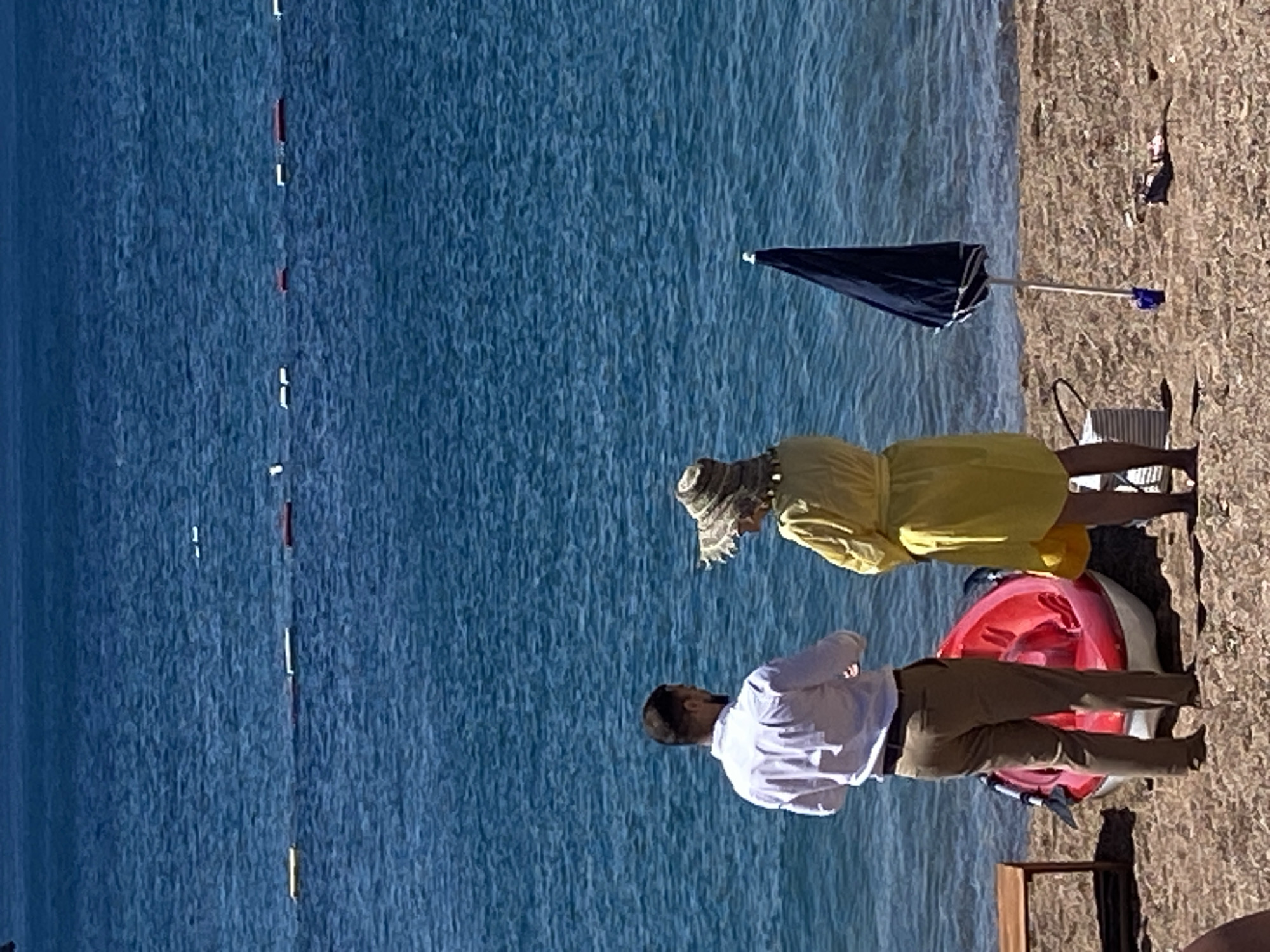 The image shows a man and a woman standing on a sandy beach facing the turquoise water in Petrovac Centar, Montenegro. The man, wearing a white long-sleeved shirt and khaki pants, stands with his back to the camera. The woman, wearing a wide-brimmed straw hat and a yellow dress, stands facing the man. Between them is a bright red kayak with white trim and a black paddle. To the right of the woman, a closed dark blue umbrella is planted in the sand. In the background, buoys mark a section of the sea. A small wooden table is visible on the left side of the frame. The scene suggests a relaxed beach setting.