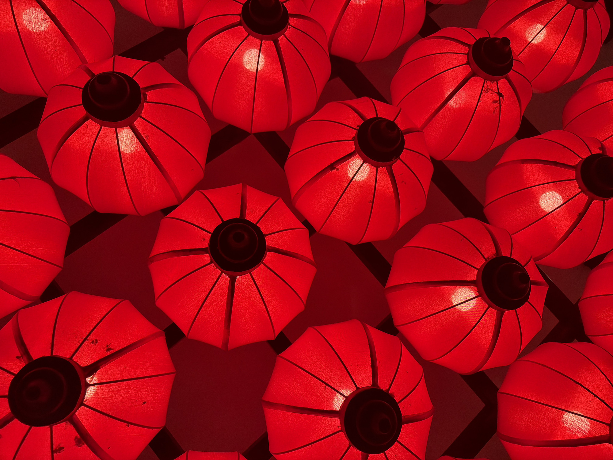 The image shows a high-angle, close-up view of numerous red lanterns. The lanterns are round with a segmented design and a black top. They are illuminated from within, casting a warm glow. A geometric pattern is visible in the background, partially obscured by the lanterns. The lanterns are densely packed. The image is monochromatic.
