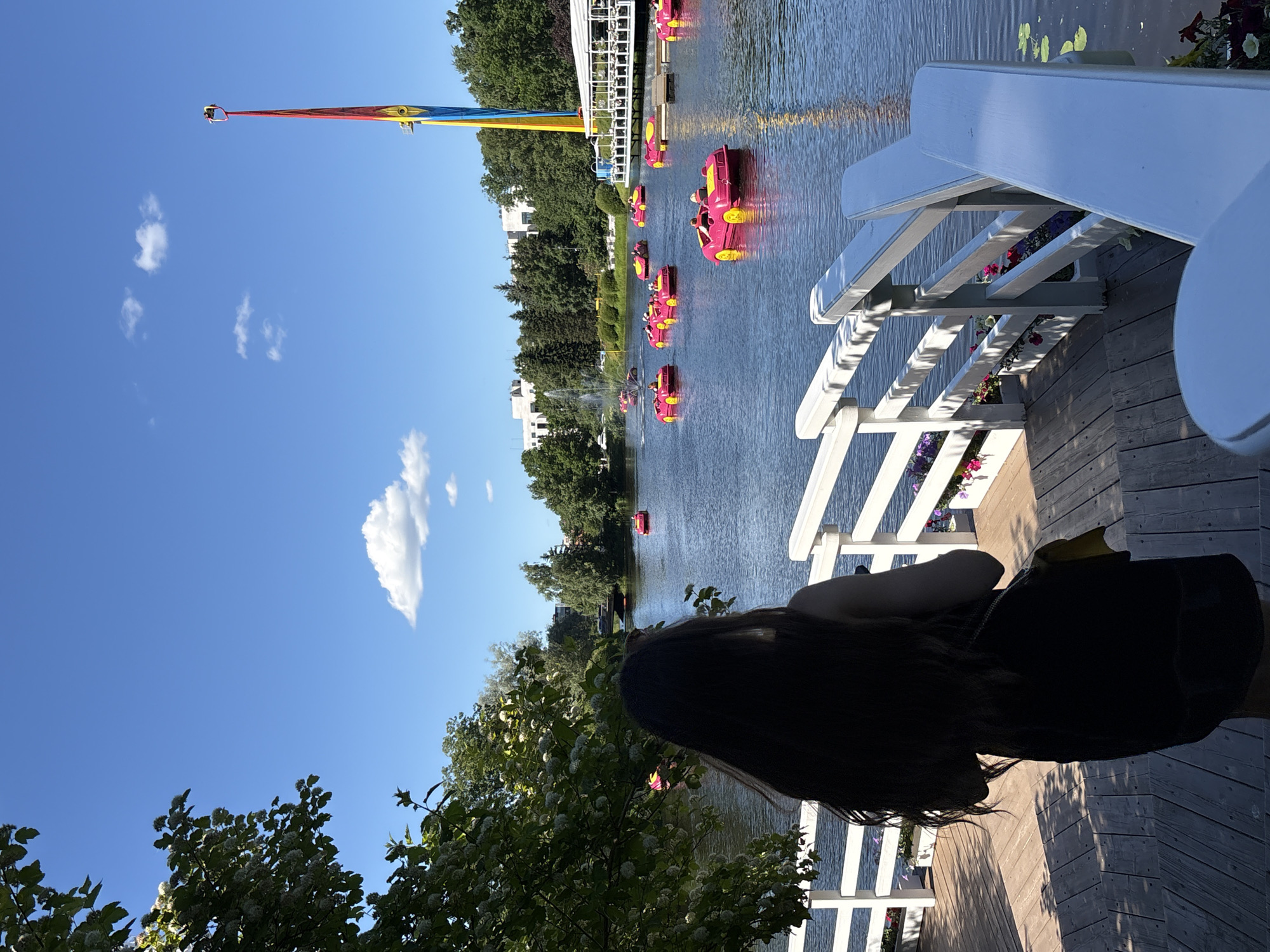 A woman stands on a wooden deck overlooking a lake filled with pink paddle boats shaped like cars. A tall, colorful amusement ride with a red, yellow, and blue tower dominates the skyline. Trees line the shore, with buildings and a fountain visible in the distance. The sky is bright blue with a few white clouds. The scene appears to be located in a park or amusement area in Saint Petersburg, Russia.