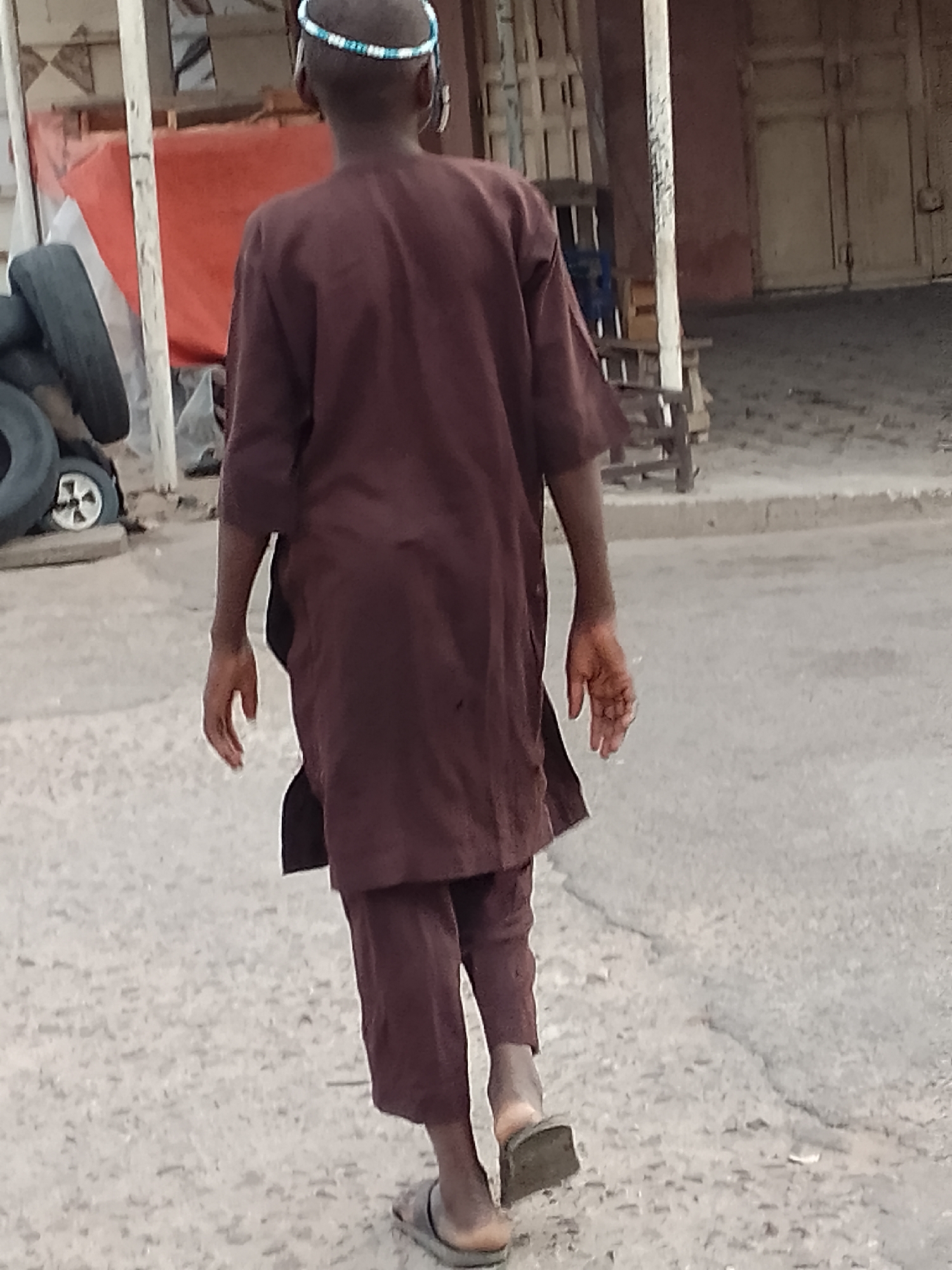 The image shows a person from behind, walking away from the camera on a dirt road in Arawa, Nigeria. The person is wearing a long, maroon tunic and matching pants, common attire in the region. They also wear a beaded headband, the beads appearing to be blue and white. On their feet, they have simple, dark-colored flip-flops.
The setting appears to be an outdoor area with some structures in the background. To the left, there are stacks of tires and a reddish-orange tarp, possibly indicating a storage or work area. A white vertical pole, likely part of a makeshift structure, is also visible. Further back, there are buildings with doorways and what might be shelves or stacked items. The ground is a dusty, uneven surface, typical of many areas in Nigeria.
The lighting suggests it is daytime, with no strong shadows indicating either very early morning or late afternoon. The weather seems to be clear. The overall scene gives a sense of everyday life in a semi-urban or rural environment. No specific emotions are evident due to the perspective. There is no visible text in the image.