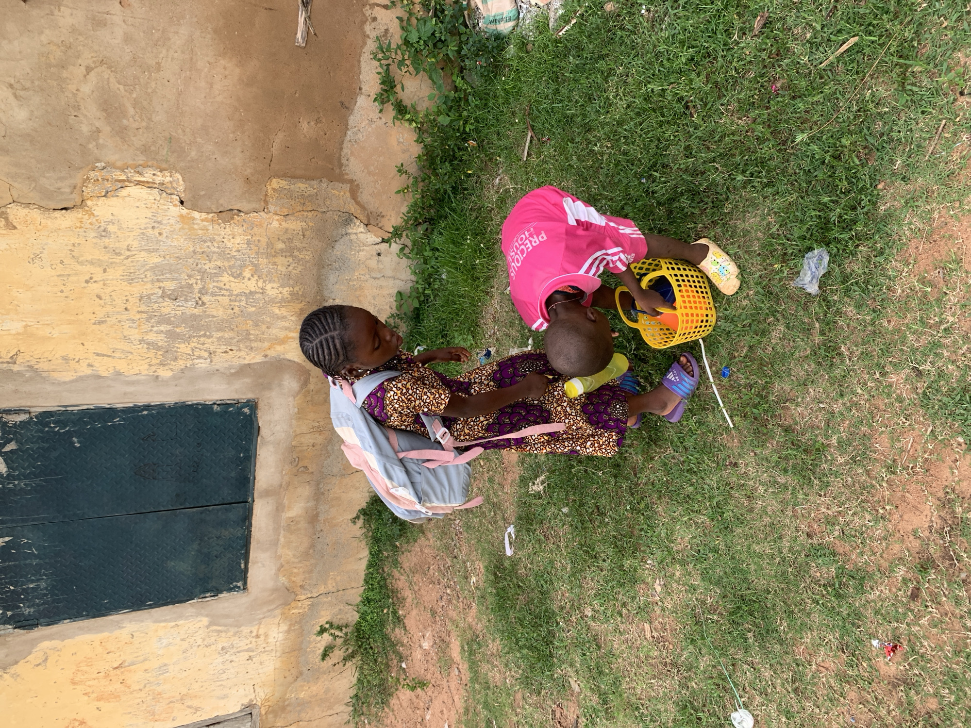 In Kabba, Nigeria, two young children are interacting outdoors next to a weathered, light-colored building with a dark metal-covered window. The child on the left, a girl with braided hair, wears a patterned dress and a light-colored backpack. She is looking down towards the other child. The child on the right, wearing a bright pink t-shirt with white lettering and yellow shoes, is bending over a yellow plastic basket, appearing to be placing items inside. The ground is covered with green grass and patches of dry earth, with some scattered debris including plastic bags and wrappers.