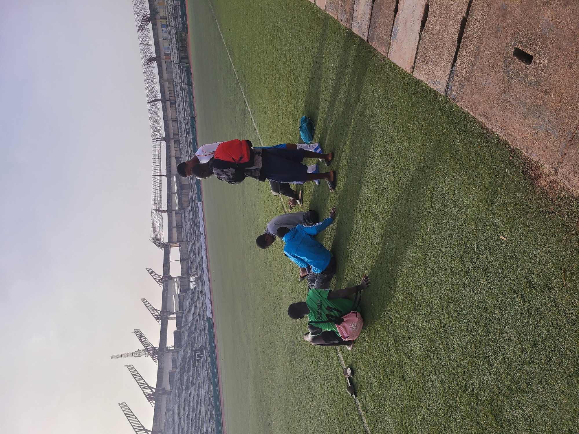 The image depicts four individuals on an artificial turf field, seemingly engaged in stretching or resting. A man wearing a white shirt with a red backpack stands with his back bent. Next to him is a man wearing a dark jacket and shorts. The third individual is wearing a blue jacket and seems to be stretching towards the ground. The fourth person, seated on the field, is wearing a green shirt and has a pink bag next to him. A pair of shoes is placed on the grass nearby. The background reveals a stadium structure, identifiable as the New Jos Stadium in Nigeria. The stadium seating is visible. The sky is lightly overcast.