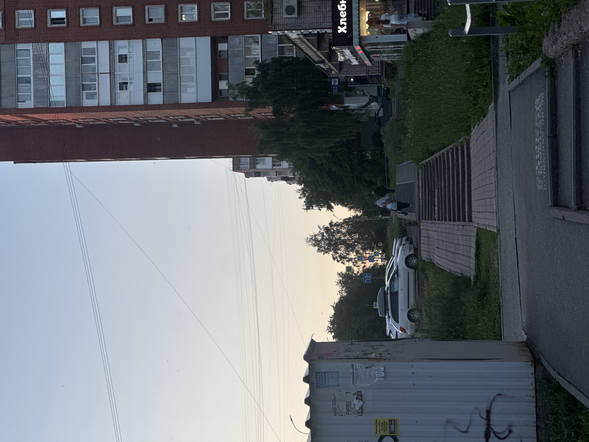 The image depicts an urban street scene in Izhevsk, Russia, under a clear, pale blue sky transitioning to a light orange-yellow hue, indicative of dusk or dawn. Numerous power lines are visible crossing the sky.
On the far left, a corrugated metal utility box or kiosk is present, covered in multiple faded and torn posters primarily in Russian. discernible texts include "РАБОТА" (Work), "СРОЧНО ТРЕБУЕТСЯ" (Urgently Required) with associated phone numbers (e.g., 8-922-517-04-94, 8-912-856-11-94, 8-912-441-31-51), "МАРКЕ ПИТАНИЕ ГАЛАНТ" (Galant Food Market), and "СТРОИТЕЛЬСТВО И РЕМОНТ" (Construction and Repair). Dark graffiti is present on the lower left of the box.
A paved asphalt path leads from the lower left foreground, curving towards the center and connecting to a series of concrete steps. To the right of this path, a wider set of concrete stairs ascends, bordered by a grassy slope on its right. Further up the slope, a blue sign with the "ПОЧТА РОССИИ" (Russian Post) emblem is visible on a pole, near a birch tree.
In the midground, several parked vehicles are present, including a white station wagon equipped with a black roof box. Beyond the parked cars and stairs, various trees with green foliage are visible. In the background right, multiple blue road signs with white arrows indicate directional turns (left, straight, right), alongside a yellow diamond sign signifying a priority road.
The right side of the image is dominated by a tall, multi-story apartment building constructed of red brick with grey concrete panels between window levels. The building features numerous windows, some with enclosed balconies, and external air conditioning units. At its base, a commercial frontage with illuminated windows displays a prominent red sign with white outlining, bearing the Cyrillic text "Хлеб" (Bread).
Several individuals are present: a person in a white top and light pants walks up the steps towards the "Хлеб" store on the far right. Another person in dark clothing ascends stairs in the midground. On the lower set of steps to the left, a woman in a blue top and light pants descends while carrying a bag, followed by another person in dark attire. Other multi-story buildings are visible in the distance to the left of the main brick structure. The ground consists of asphalt, paving stones, and areas of green grass.