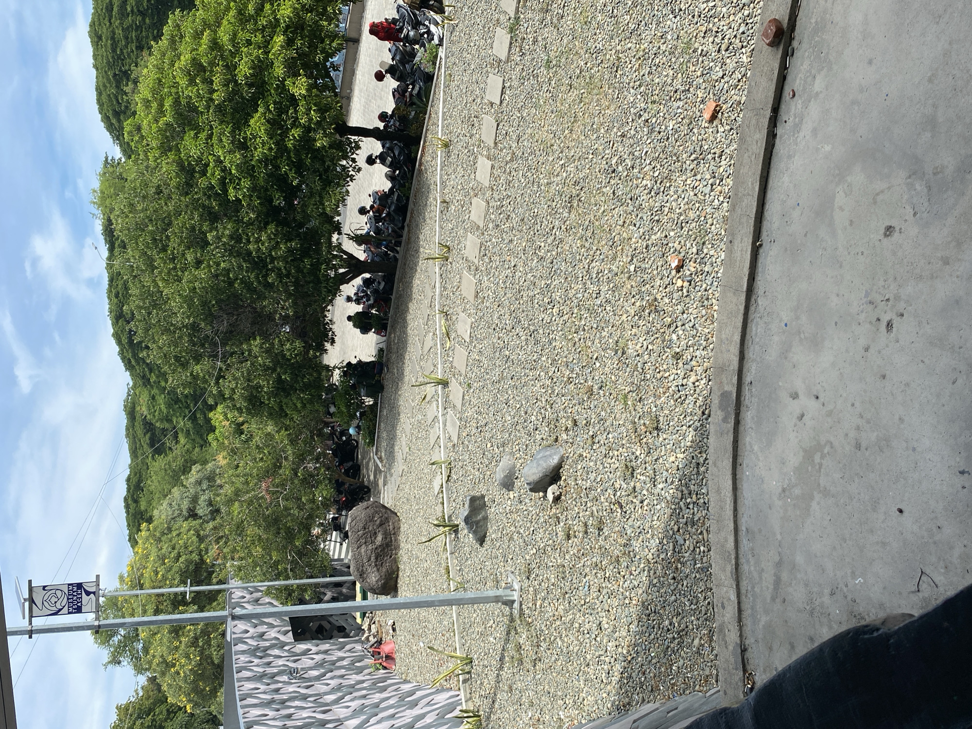 The image depicts an outdoor scene in Banda Aceh, Indonesia, likely at the Museum Tsunami Aceh given the banner on the pole. The foreground shows a concrete walkway adjacent to a gravel-covered area with several large rocks and stepping stones arranged in a path. A tall metal pole, possibly supporting a banner and solar panel, stands in the gravel. In the background, numerous parked motorcycles are visible under the shade of lush green trees. A patterned wall, potentially part of the museum's exterior, is on the left side of the frame. The sky is blue with scattered clouds.