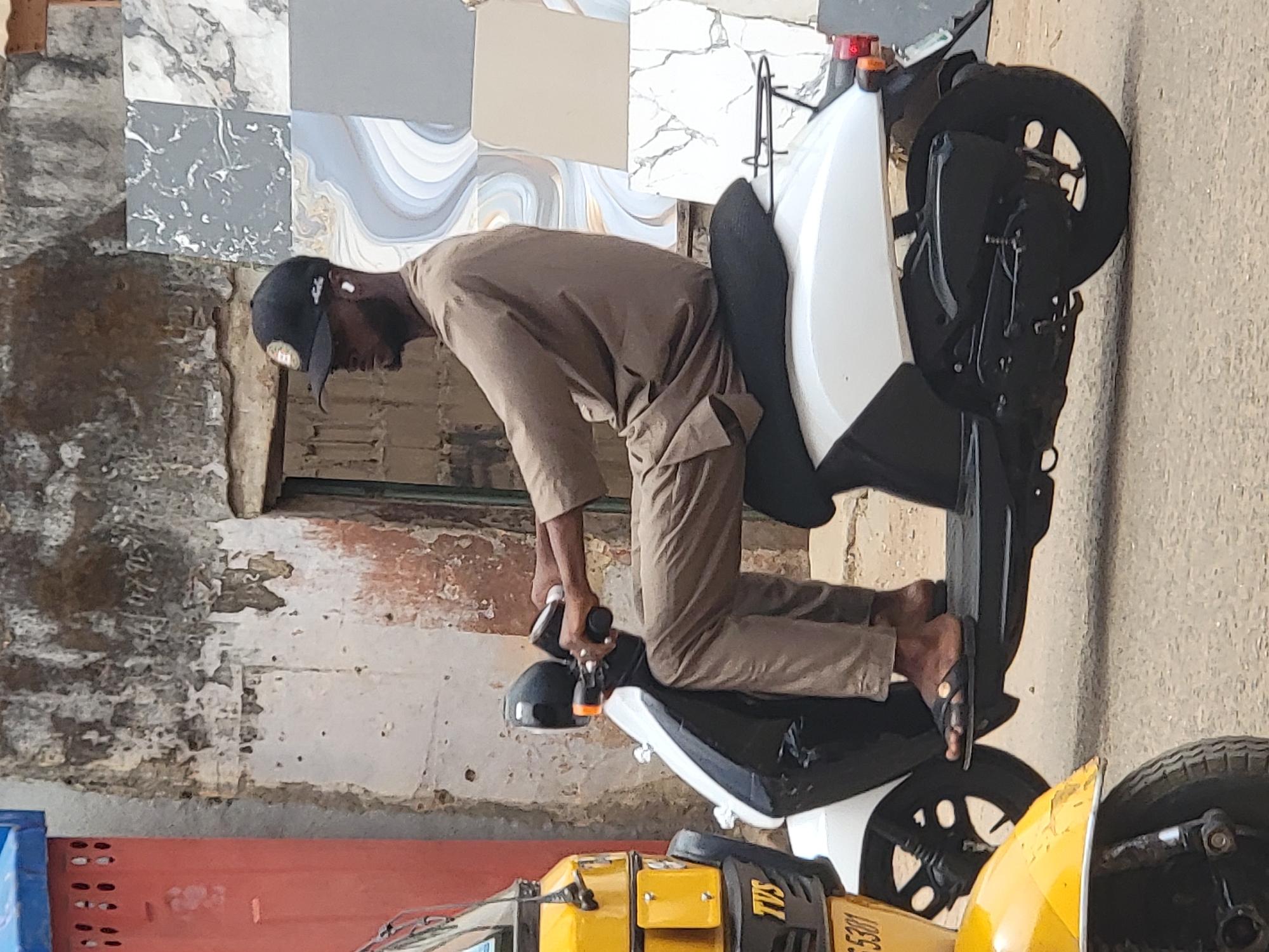 A man wearing a brown shirt, trousers, cap, and sandals is sitting on a white and black motorcycle. He is holding the motorcycle's handlebars. The background consists of a building with grey and white tiling on the upper portion and peeling paint on the lower portion. Other parts of a yellow and black motorcycle are visible at the bottom of the frame. The location is Maiburiji, Nigeria.