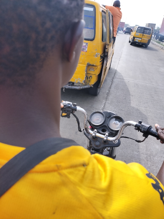 The image depicts a first-person perspective from the back of an individual operating a motorcycle on a multi-lane asphalt road in Alaiyabiagba, Nigeria. The motorcycle operator, whose dark-skinned head and right shoulder are visible, wears a bright yellow shirt with a black strap traversing the right shoulder. Their right hand is positioned on the motorcycle's right handlebar, which features control switches. The motorcycle's instrument panel, including two circular gauges, is visible between the chrome handlebars.
Immediately to the right of the motorcycle, a yellow commercial minibus (commonly known as a "danfo" in Nigeria) is either stopped or moving slowly. The minibus exhibits significant wear, including visible damage to its rear bumper and body panels. Its side sliding door is open, and an adult male, clad in an orange shirt, is partially visible, leaning out or in through the open doorway, holding onto the door frame or roof.
Further down the road, several other vehicles are present, including at least one additional yellow minibus and multiple larger trucks, suggesting moderate to heavy traffic conditions. The sky above is light, indicating an outdoor, daytime scene.