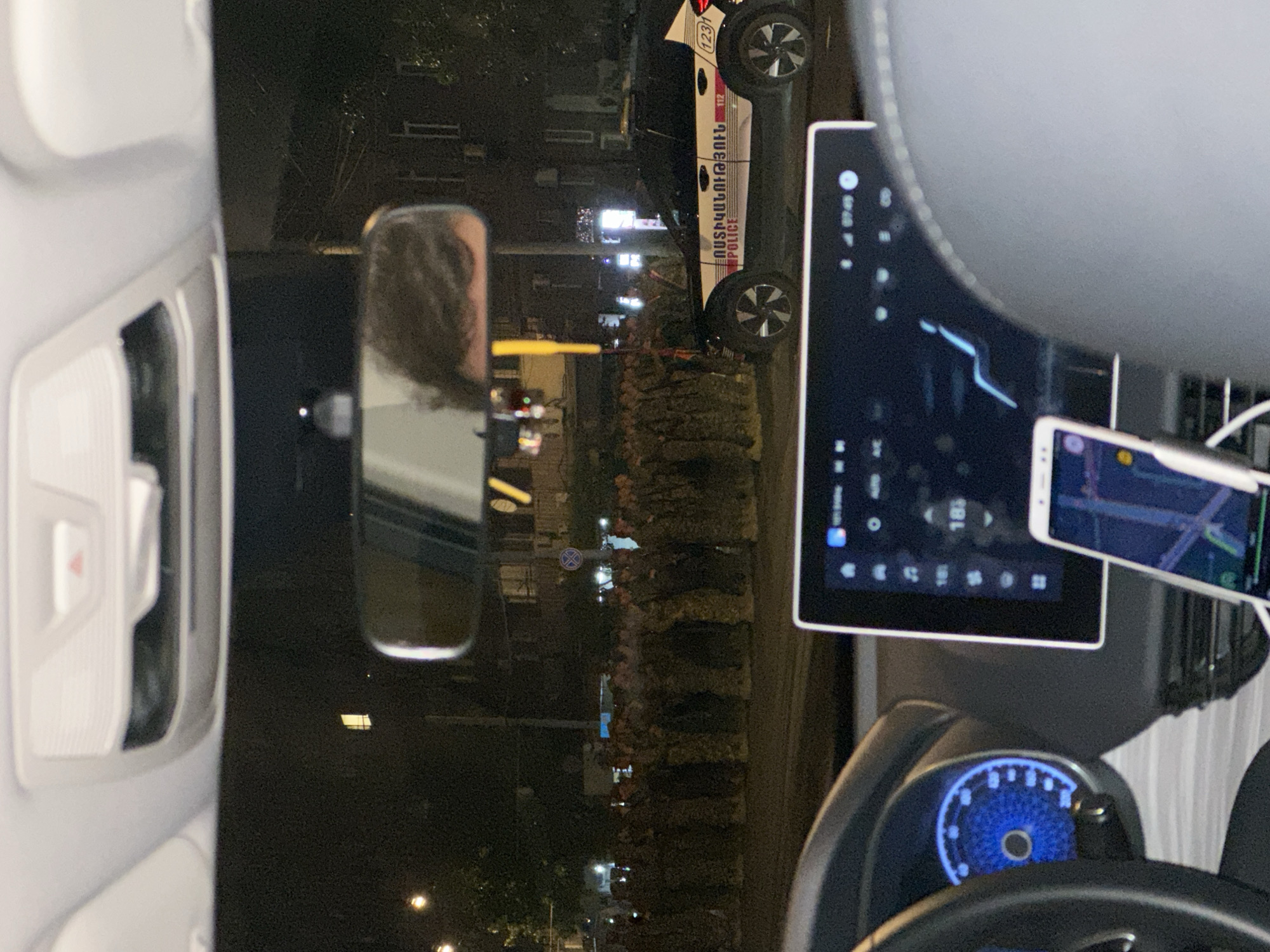 The image shows the interior of a car at night, likely in Yerevan, Armenia, based on the presence of a police car with Armenian writing on its side in the side mirror view. The dashboard is visible, featuring a screen displaying what appears to be a navigation map. A smartphone is mounted next to the screen, also showing a navigation map. The driver's side mirror reflects the street scene, including streetlights, buildings, and the Armenian police car. The scene suggests the car is stopped or moving slowly, possibly observing or following the police vehicle. The interior is dark, illuminated primarily by the dashboard and phone screens.