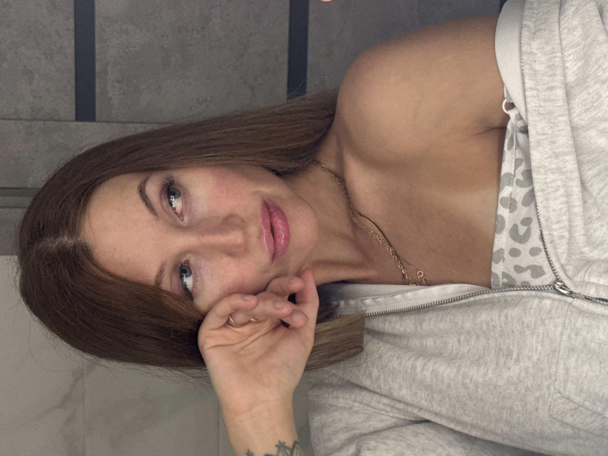 A woman with light brown hair and blue eyes is looking at the camera with her hand partially covering her face. She is wearing a light grey zip-up hoodie and a gold necklace. The background appears to be a grey tiled wall. There is a tattoo visible on her wrist. No other information is available to place the scene in Chaikovsky, Russia.