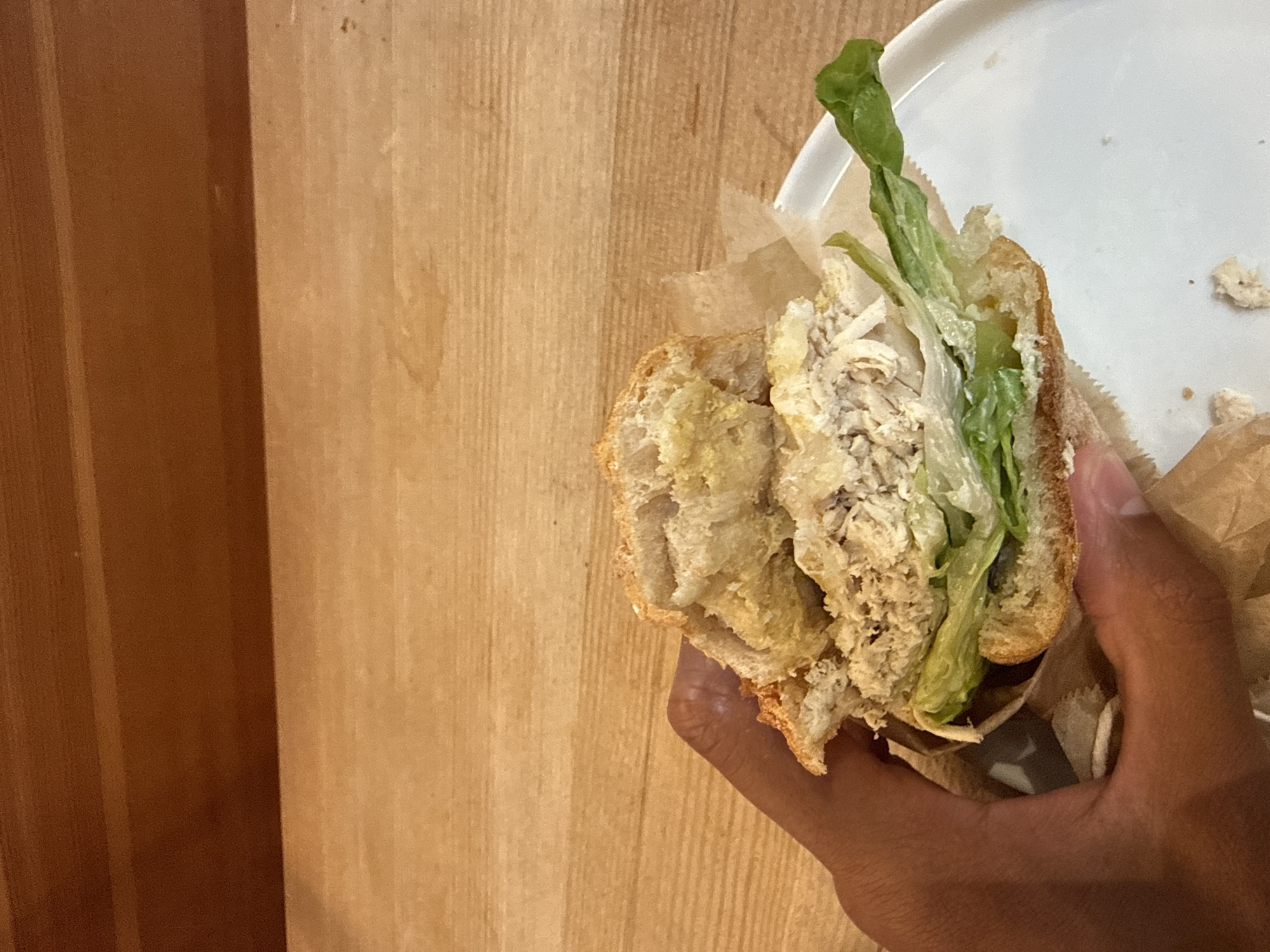 The image presents a close-up, vertical shot of a hand holding a sandwich against a wooden backdrop. A hand with a dark skin tone firmly grips a large sandwich, likely a sub or baguette, filled with what appears to be shredded chicken salad or tuna salad, generously layered with crisp green lettuce. The bread is a light, rustic-style roll, showing a textured crust and soft interior.
The sandwich is partially encased in light brown paper or wax paper, suggesting it is either a freshly prepared meal or a takeaway item. To the upper right, a segment of a white ceramic plate is visible, bearing a few crumbs, which might indicate partial consumption of the sandwich.
The background consists of a wooden surface, divided into two prominent vertical sections. The left section features a darker, reddish-brown wood panel, while the right section displays a lighter, golden-brown wood panel, both exhibiting distinct vertical grain patterns. The lighting suggests an indoor setting, likely ambient. The activity captured is a person holding their meal, possibly about to eat or having already started. There is no visible text within the image, and no specific time of day, weather, or location cues are discernible beyond the indoor context.