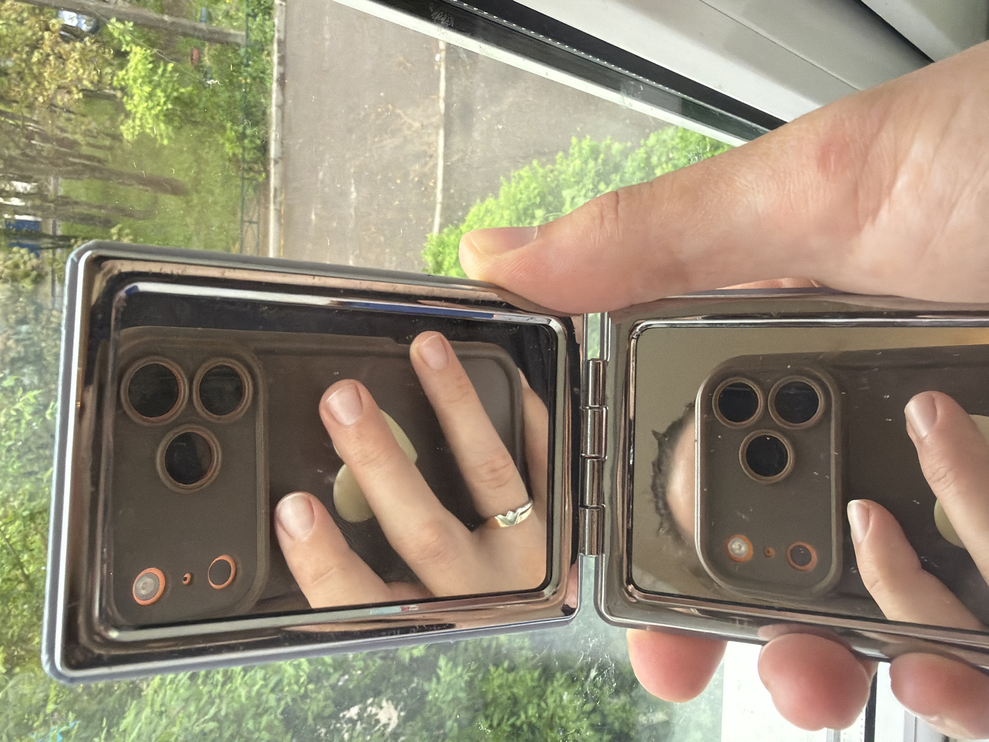 A hand holds an open, hinged compact mirror reflecting a smartphone. The smartphone has a triple-lens camera. The hand's middle finger features a ring. The background, visible through the compact mirror and behind the hand, shows greenery and a paved area, likely an outdoor environment, possibly a park or urban area. The background is also visible through a window frame. The reflection implies the person holding the mirror is taking a picture of themself and their phone. The scene appears to be indoors, given the window frame.