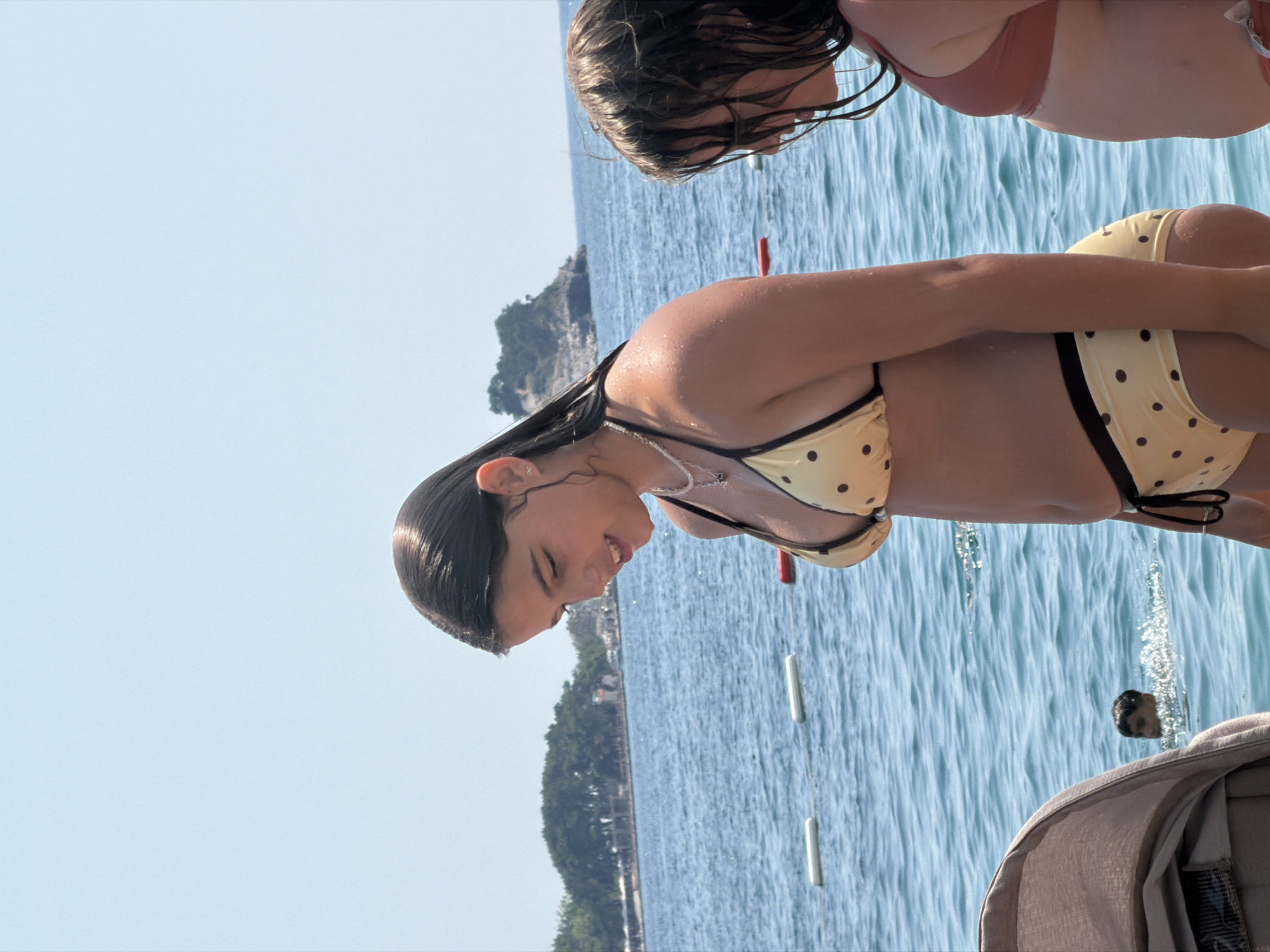 The image shows two young girls in swimsuits at a beach in Budva, Montenegro. The girl in the foreground is wearing a yellow bikini with black polka dots and has wet, dark hair. She is smiling and looking down, possibly at the water. The second girl, partially visible in the background, is wearing a pink bikini top and also has wet, dark hair. They appear to be in the ocean, with blue water and a hazy sky. There are buoys or markers in the water, and a tree-covered hillside with buildings is visible in the distance, suggesting a coastal town. The lighting and the water suggest it is daytime, likely sunny. The overall mood seems cheerful and relaxed, with the girls enjoying a day at the beach.
