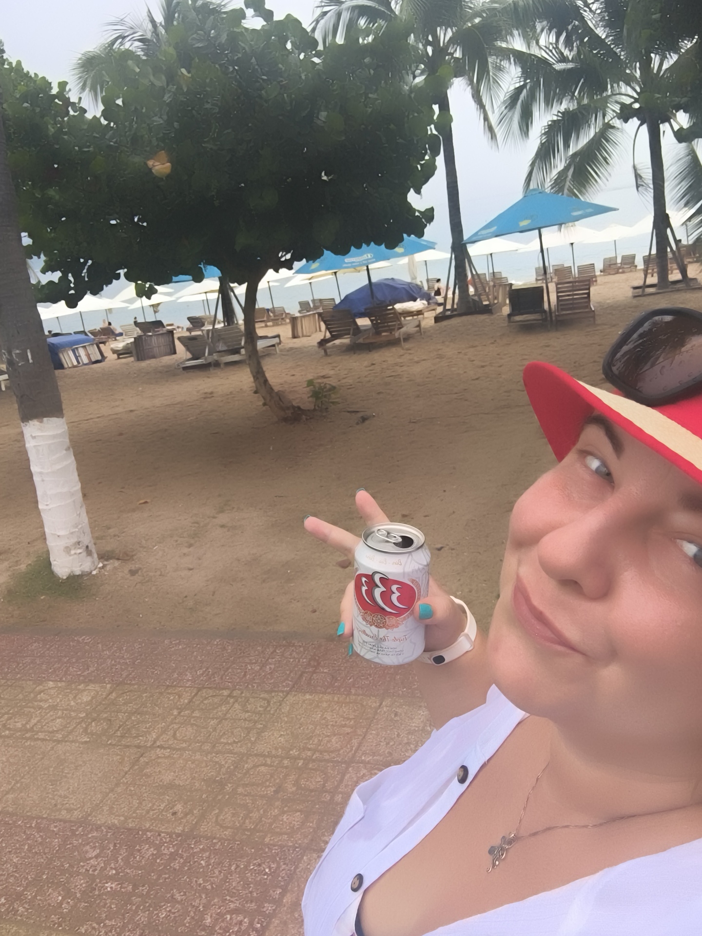 In Nha Trang, Vietnam, a woman in a white shirt and red hat is taking a selfie on a sandy beach. She is holding a can of beer and has a relaxed expression. In the background, palm trees, beach chairs, and umbrellas are visible, suggesting a tropical vacation setting. The beach appears to be somewhat cloudy.