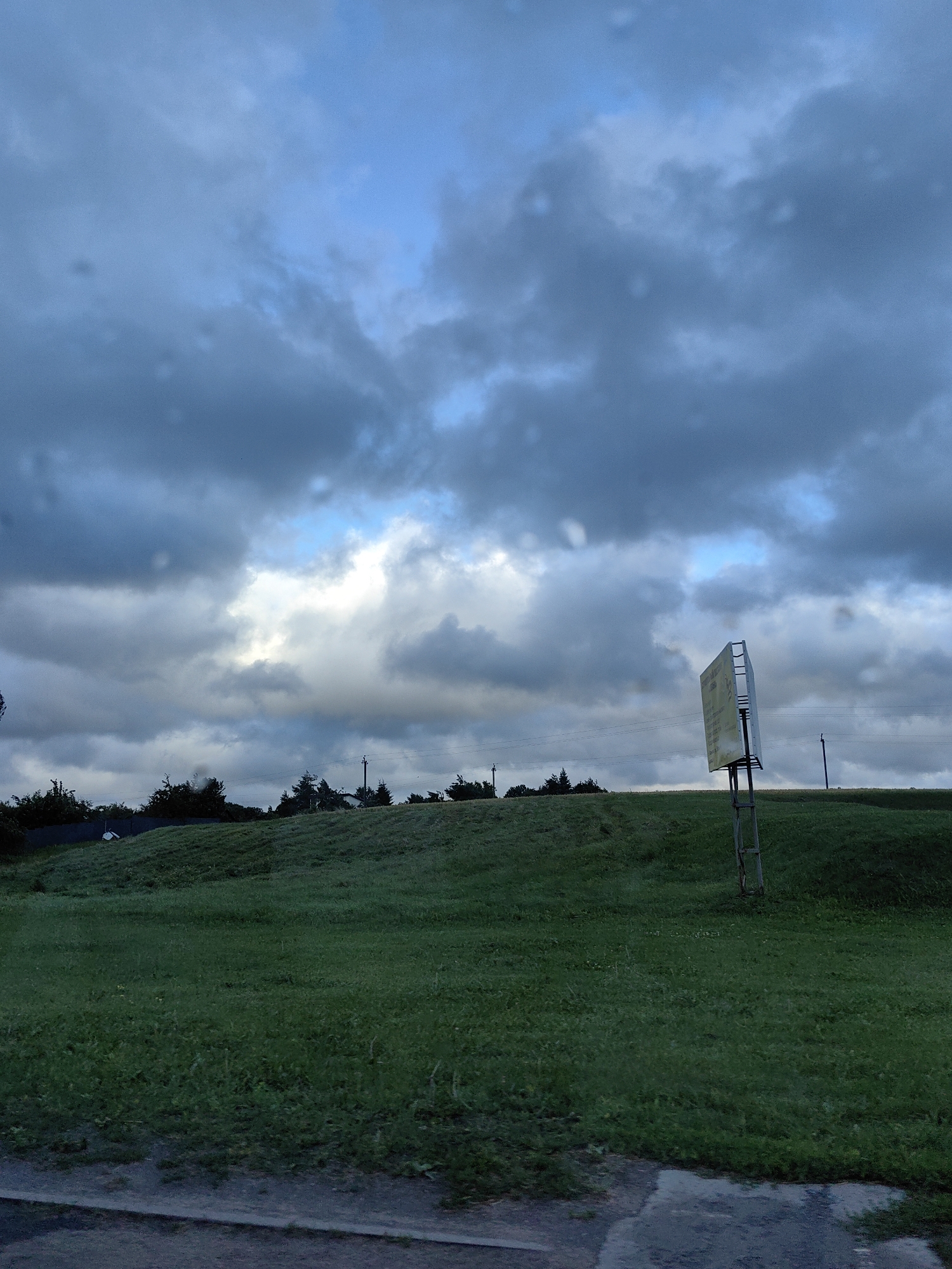 The image depicts a grassy hill under a dramatic, cloudy sky with patches of blue. On the right side of the hill, a tall, metal signpost holds a rectangular sign with yellow and white lettering. The sign is tilted and appears weathered. The hill is covered in lush green grass, and in the distance, a line of trees and utility poles are visible. The lower portion of the image shows a paved surface, possibly a road or sidewalk, with a reflective puddle. The overall atmosphere is overcast and possibly damp, with subtle indications of rain or moisture on the camera lens. The scene appears to be an outdoor landscape, likely in a rural or suburban area, possibly near a road. There are no people visible in the image. Based on the provided context, the location is identified as Фаніпальскі сельскі Савет, Belarus.