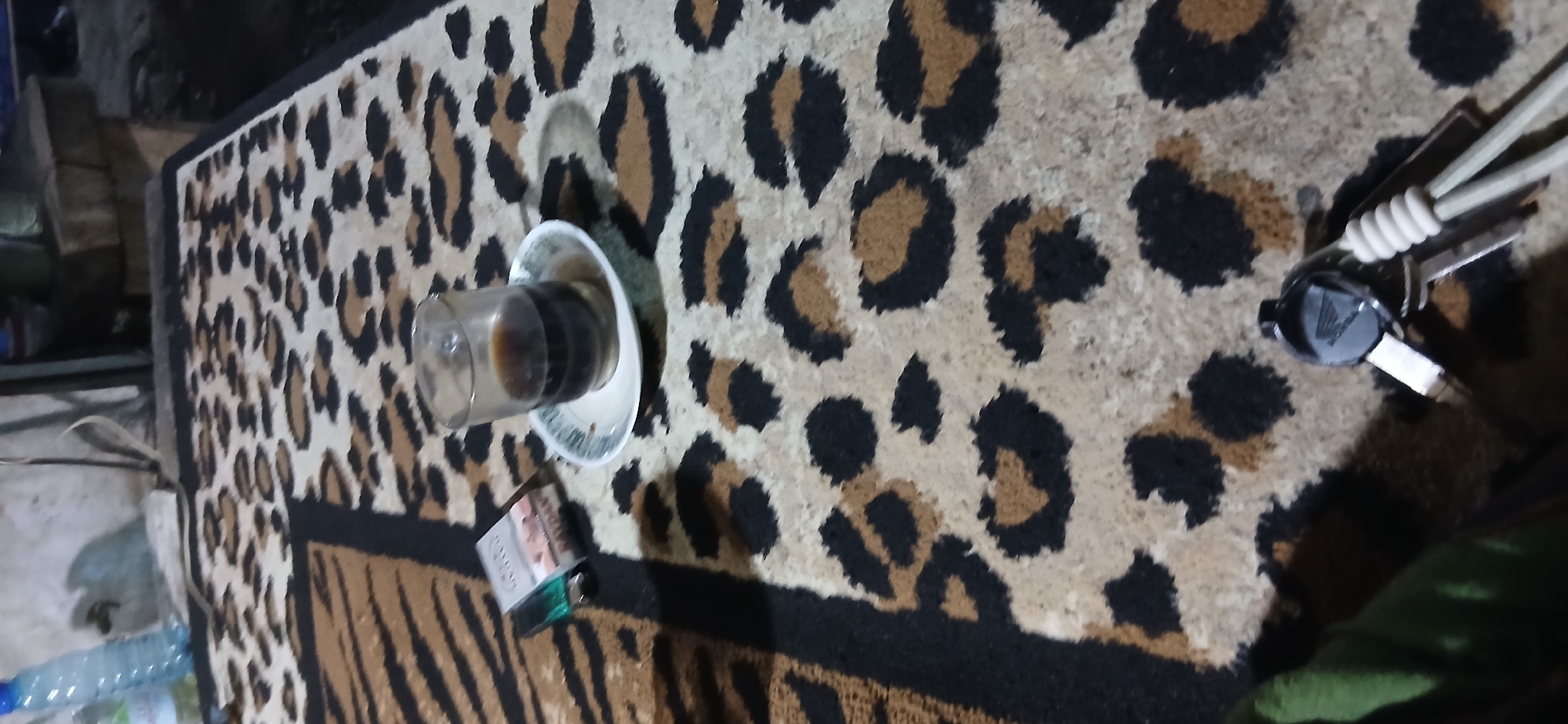 A low-angle, medium shot depicts a close-up view of a table covered with a leopard-print tablecloth. On the table, a glass of dark liquid, likely coffee, sits on a small white saucer. To the left of the glass, a pack of cigarettes, labeled "RAYDAN," is visible. In the foreground, a set of keys with a "HONDA" keychain is placed. The background is out of focus but appears to be an indoor setting with a wall and some indeterminate objects. The lighting is dim, casting shadows across the scene. The image was taken in Demangan, Indonesia.