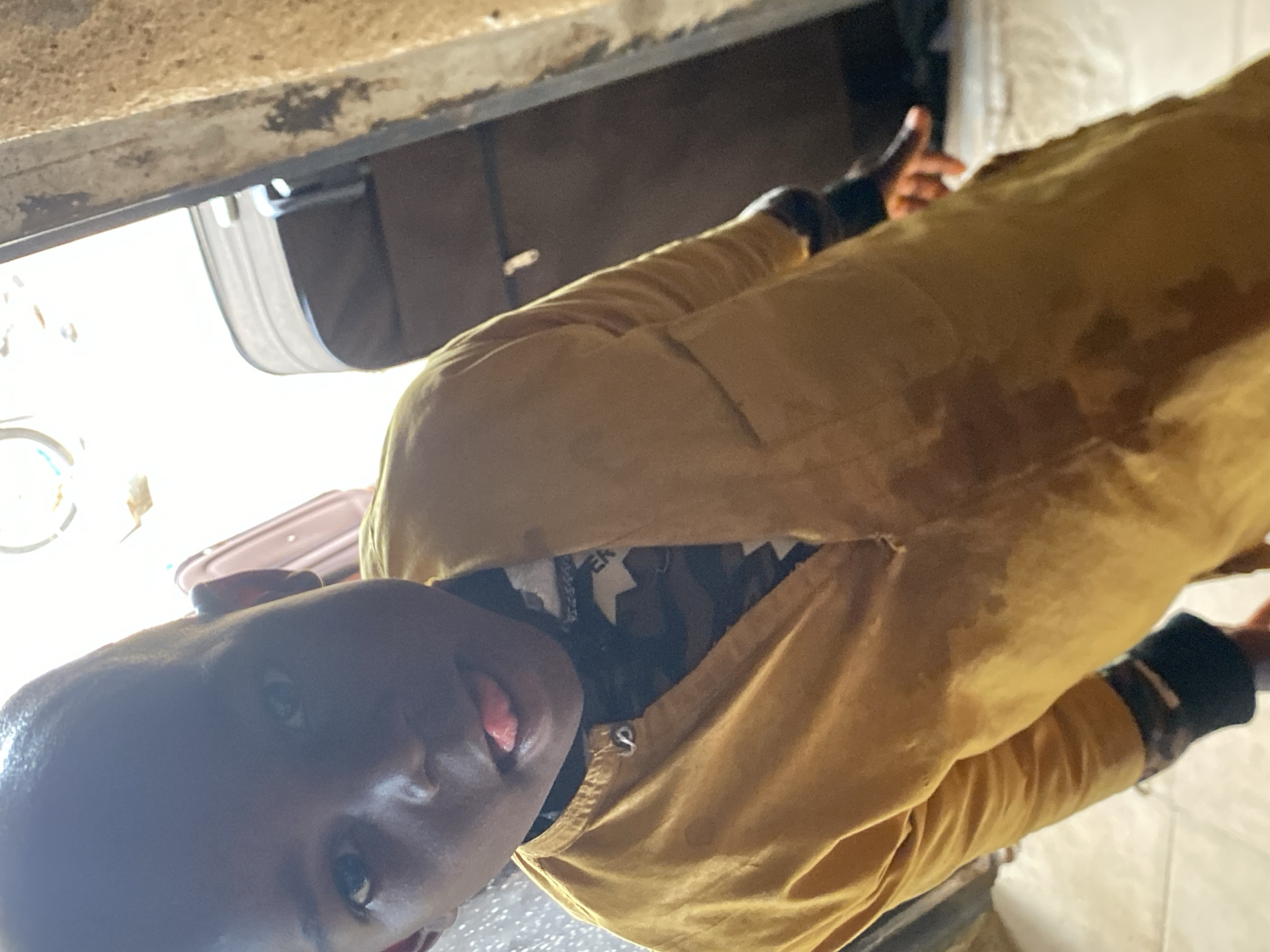 In Bukuru, Nigeria, a young boy wearing a yellow shirt with a camouflage pattern underneath sticks his tongue out at the camera, his face partially obscured by shadow. He stands in a doorway, with luggage visible in the background. The scene appears to be indoors, with natural light streaming in from the right side of the frame. The wall to the right is rough and textured, suggesting it may be made of mud or concrete.