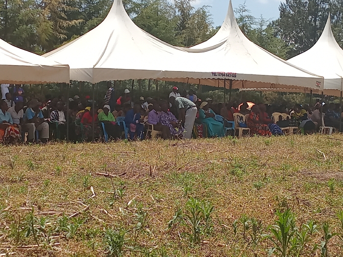 The image shows a gathering of people in Lwandeti, Kenya (latitude: 0.554, longitude: 34.829). The people are seated under several white tents, facing the viewer. There are numerous people seated in rows under the tents, with some individuals standing. Many of the people are wearing brightly colored clothing. The tents appear to be arranged in a line, with a number of trees visible in the background. The ground in front of the tents is covered in dry grass and other vegetation. There is a sign on one of the tents that reads "Tel: 0744 643 622."